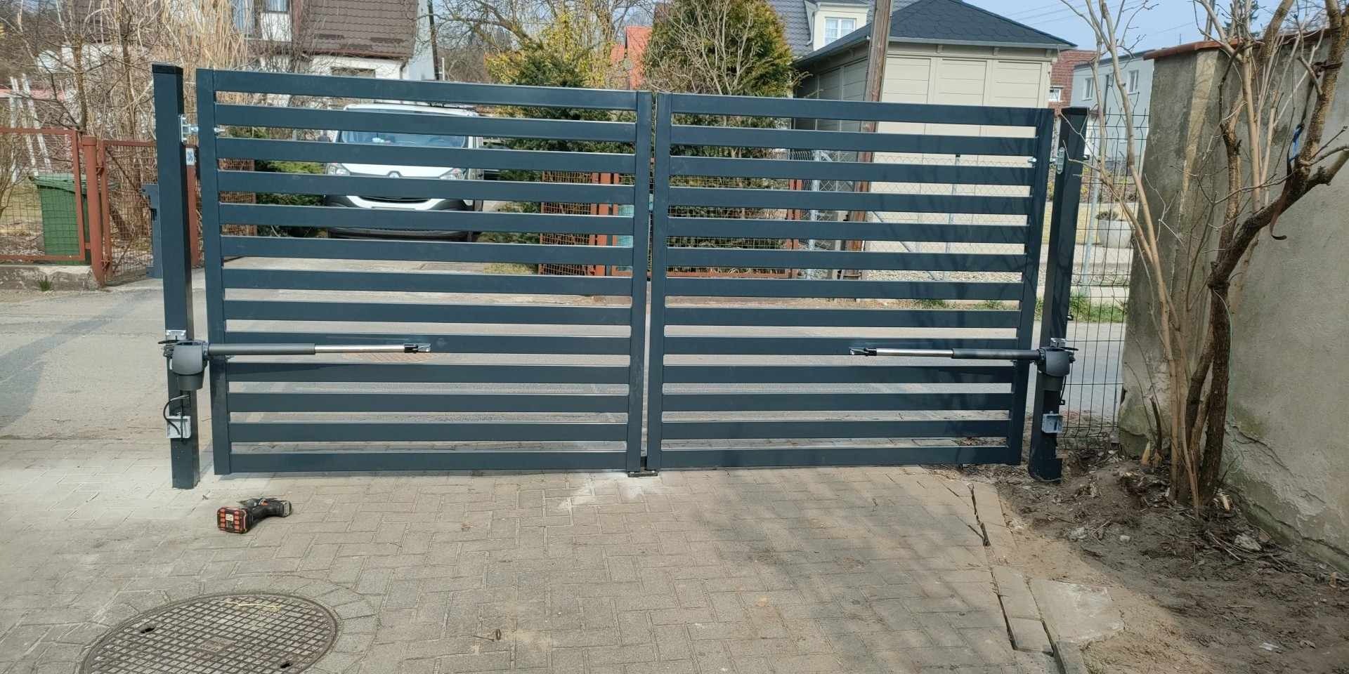 Nowoczesna, pozioma brama dwuskrzydłowa w kolorze grafitowym z zamontowaną automatyką, na pierwszym planie wkrętarka akumulatorowa