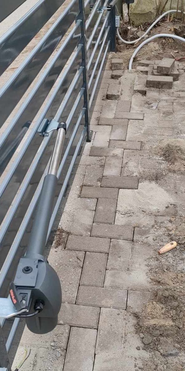 Szara brama z widocznym siłownikiem, kostka brukowa wokół w trakcie układania, widoczne przewody elektryczne.