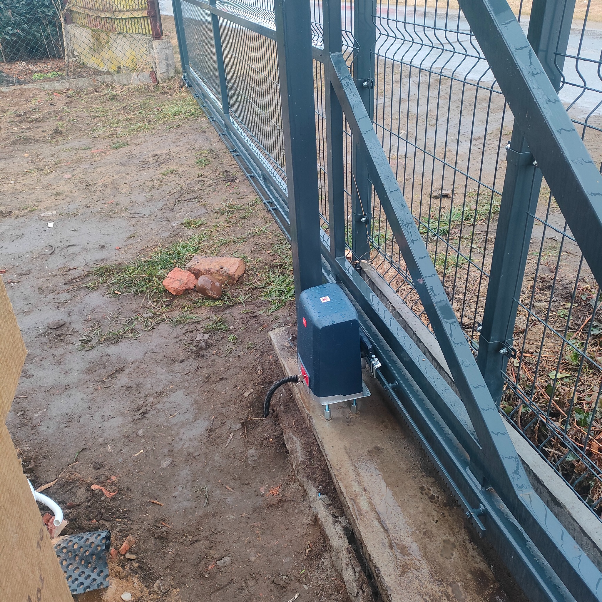 Szara, przesuwna brama panelowa z widocznym napędem elektrycznym zamontowanym na betonowej podstawie. Na ziemi błoto i fragmenty cegieł.