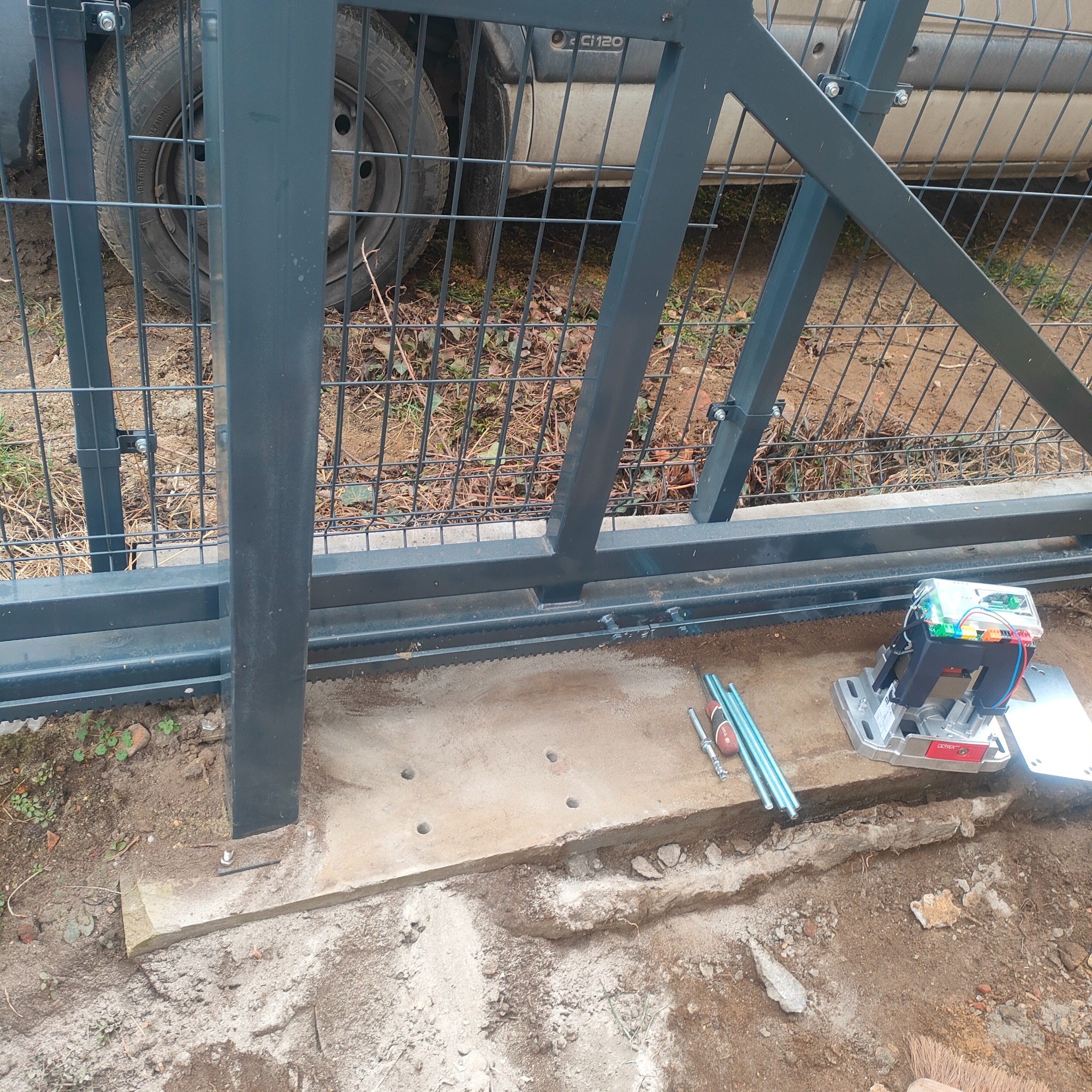 Montaż szarej bramy przesuwnej: widoczny fundament z wywierconymi otworami, narzędzia i automat do bramy Ultra na tle zaparkowanego samochodu.