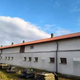 Długi budynek gospodarczy z nowym dachem z czerwonej dachówki, widoczne kominy i rynny, przy ścianie sterty starych płyt dachowych i materiałów budowlanych.