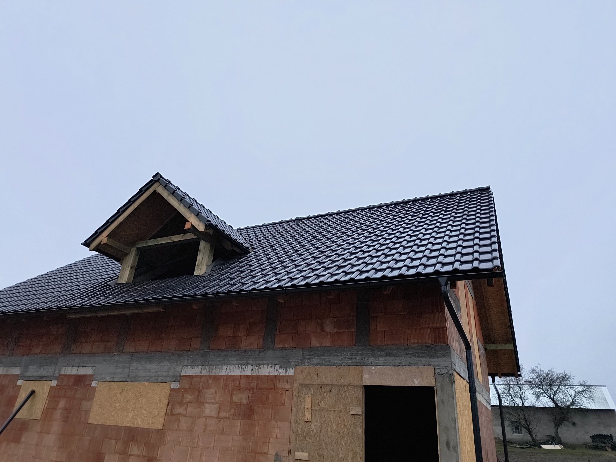 Dom w stanie surowym z czarnym, błyszczącym dachem i rynnami, widoczny fragment poddasza z drewnianą konstrukcją, nieotynkowana ściana z czerwonej cegły.
