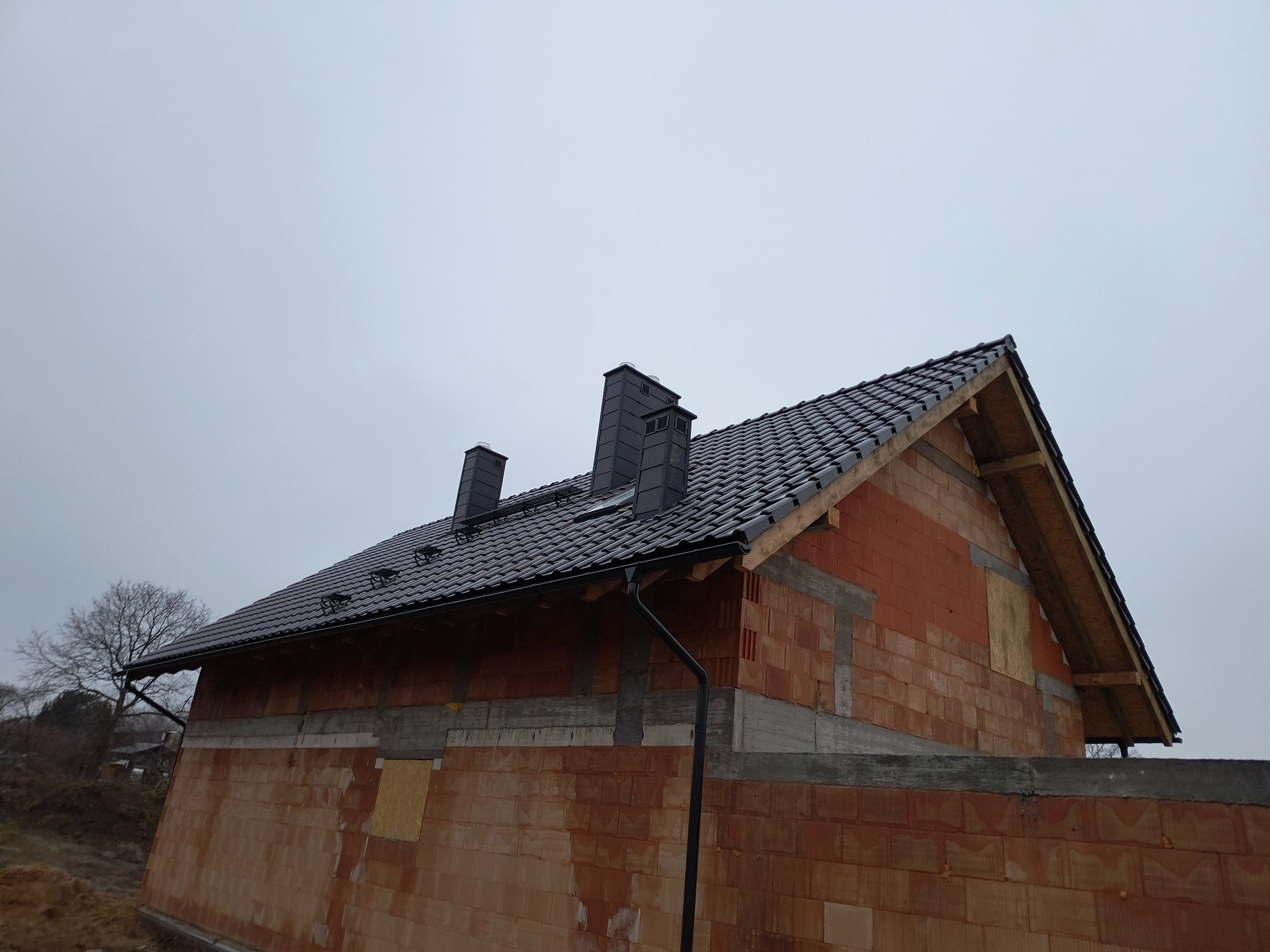 Surowy stan budynku z ciemną dachówką i rynnami, widoczne kominy i niezakończona elewacja z czerwonej cegły