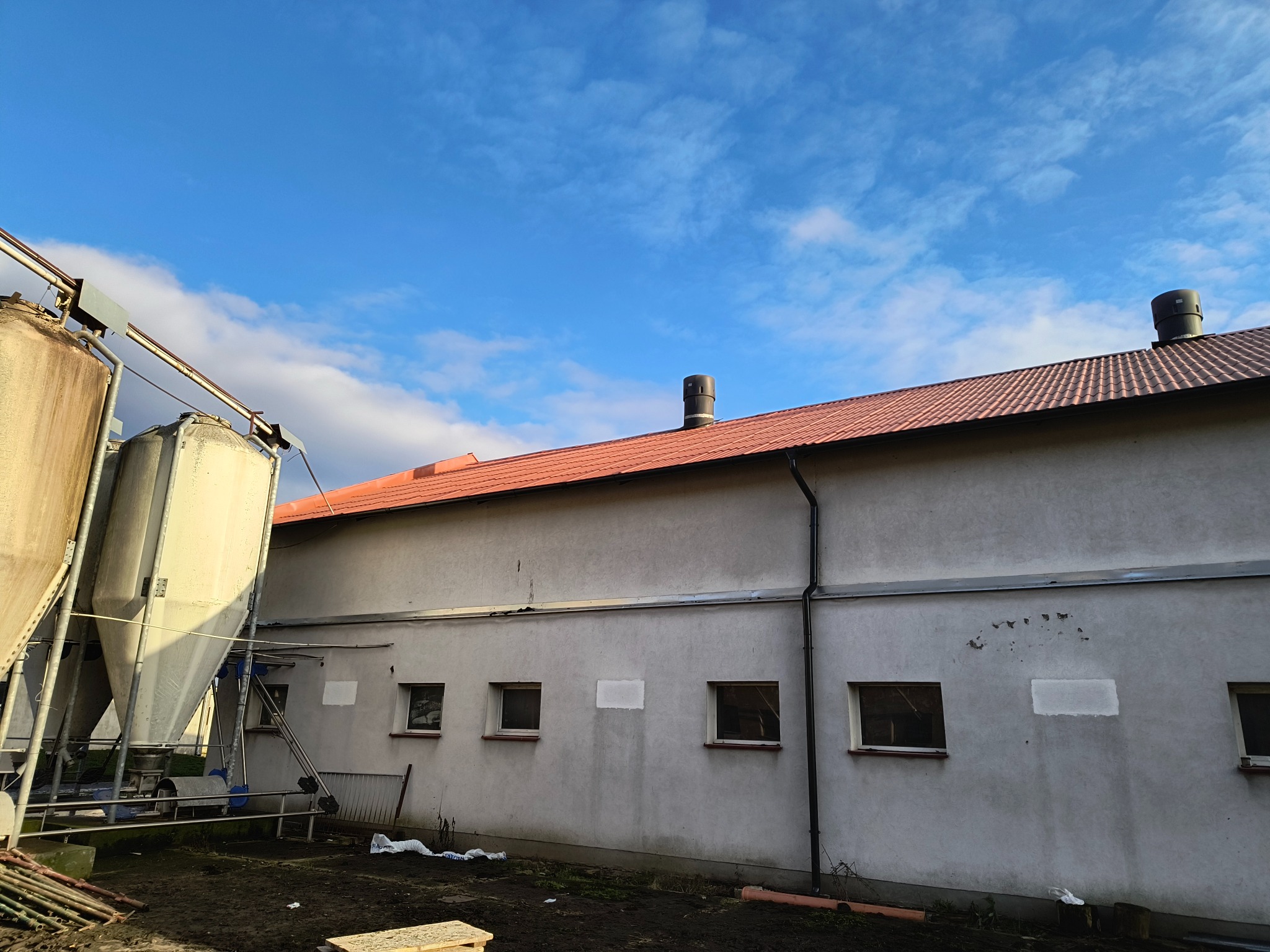 Budynek gospodarczy z czerwoną dachówką, widoczne silosy i rynna spustowa na tle błękitnego nieba z chmurami.