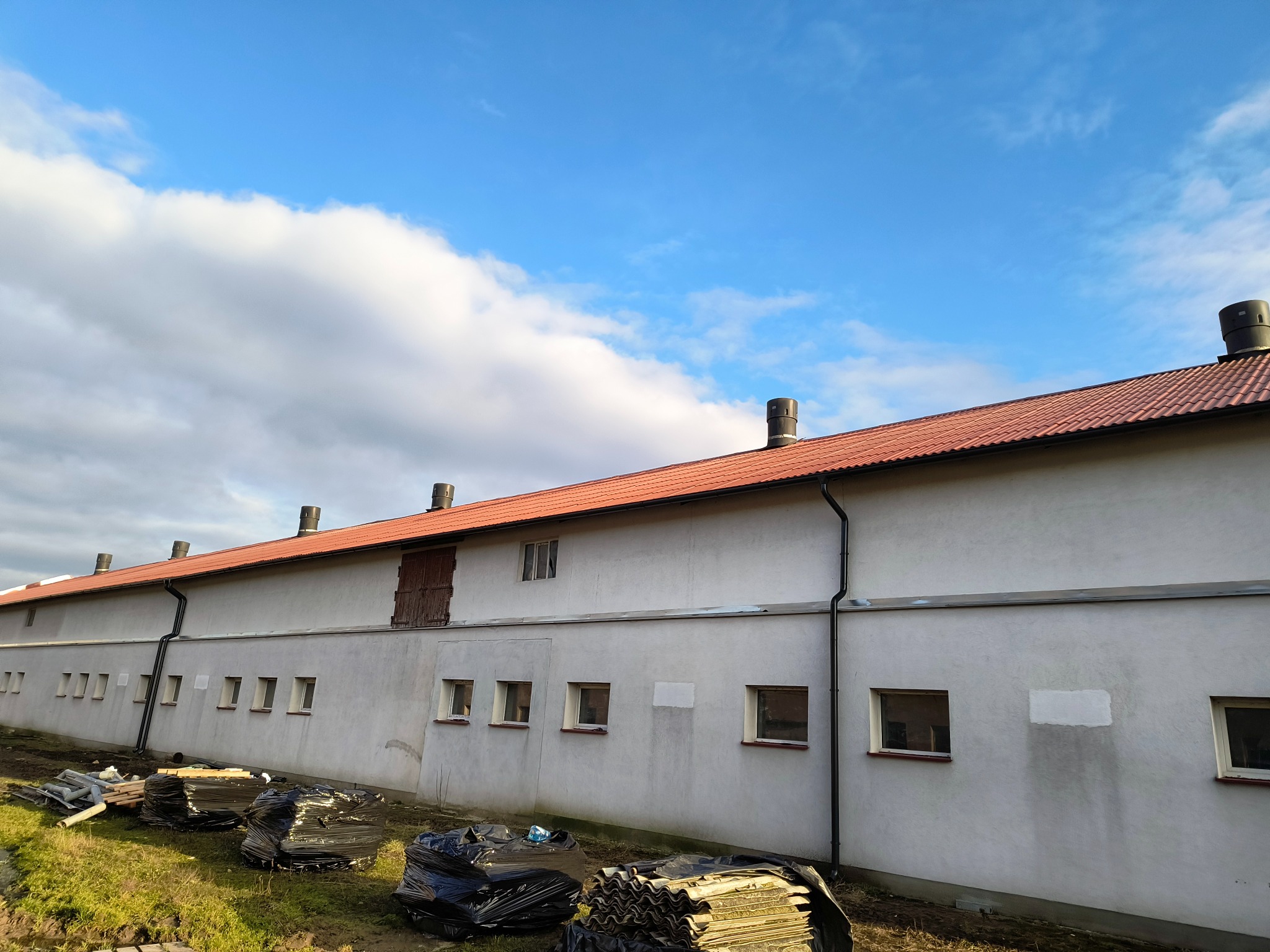 Długi budynek gospodarczy z nowym dachem z czerwonej dachówki, widoczne kominy i rynny, przy ścianie sterty starych płyt dachowych i materiałów budowlanych.