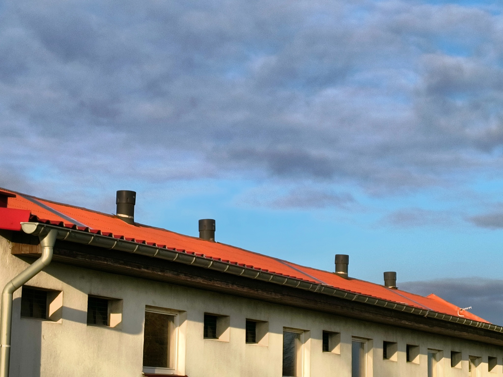 Pomarańczowy dach z czterema kominami na tle błękitnego nieba z chmurami, widoczne rynny i okna budynku.