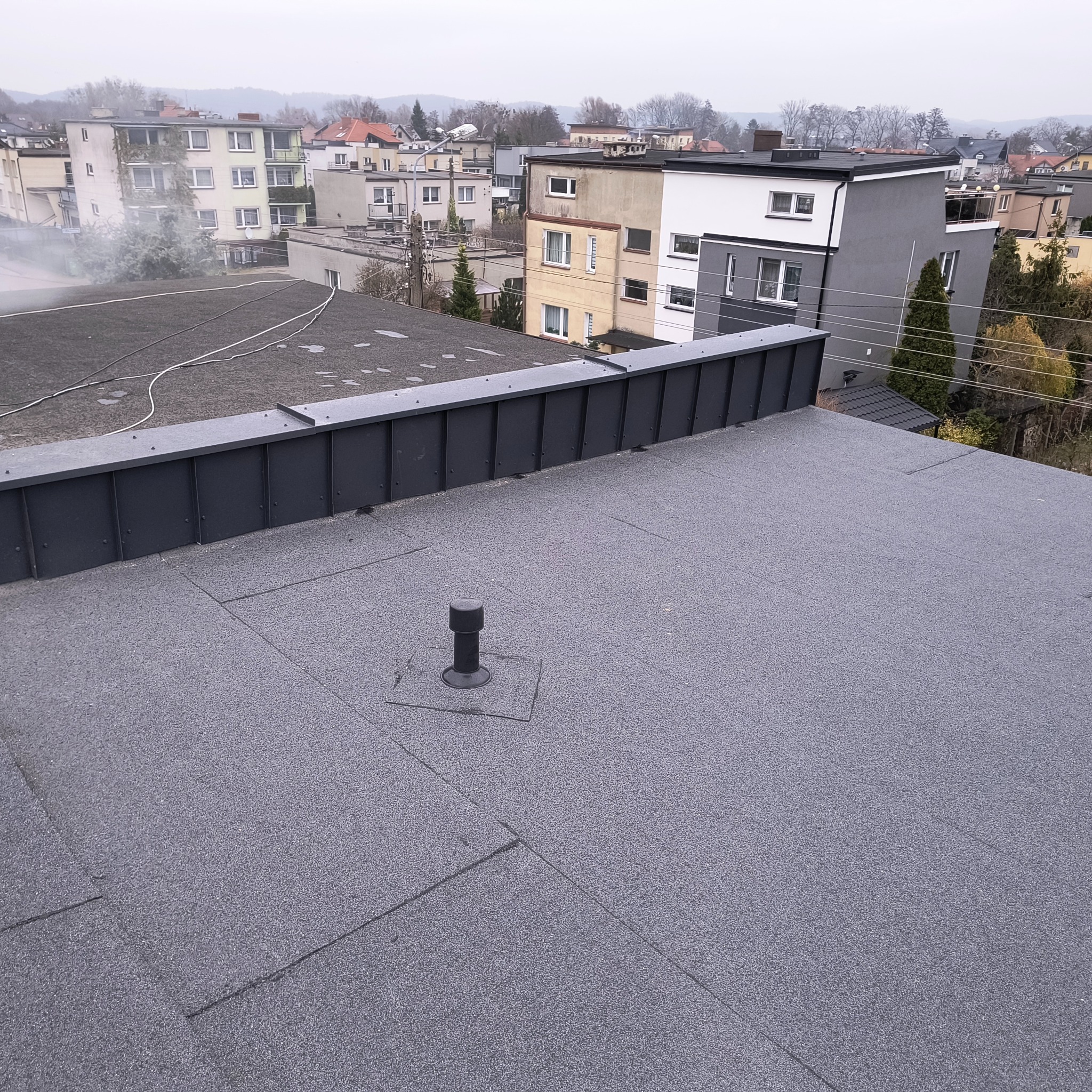 Płaski dach pokryty papą termozgrzewalną z widocznym kominkiem wentylacyjnym i obróbką blacharską na krawędzi, w tle zabudowa miejska w pochmurny dzień.