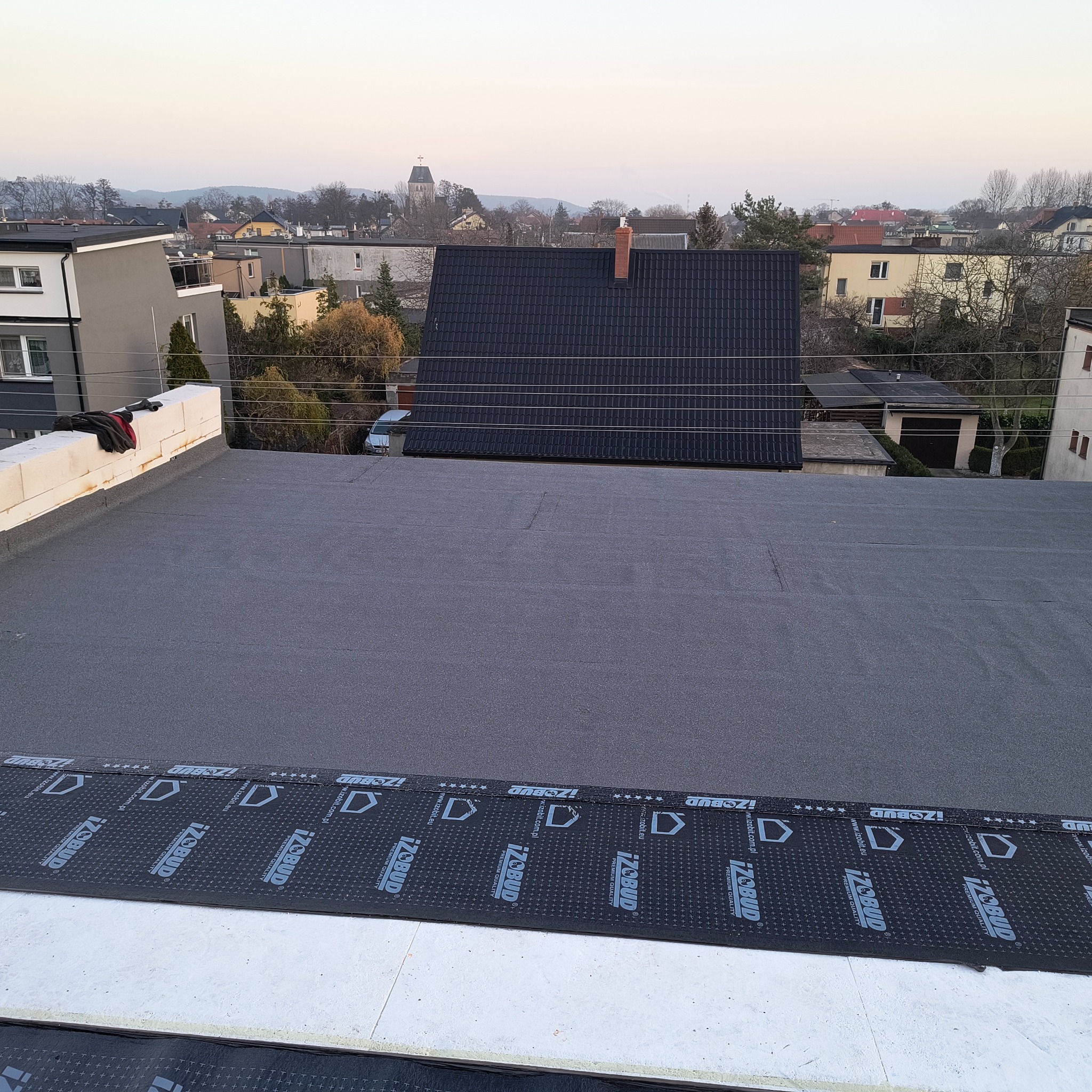 Widok z góry na płaski dach pokryty papą z widocznymi fragmentami izolacji marki Izobud, w tle zabudowa miejska i kościelna wieża.