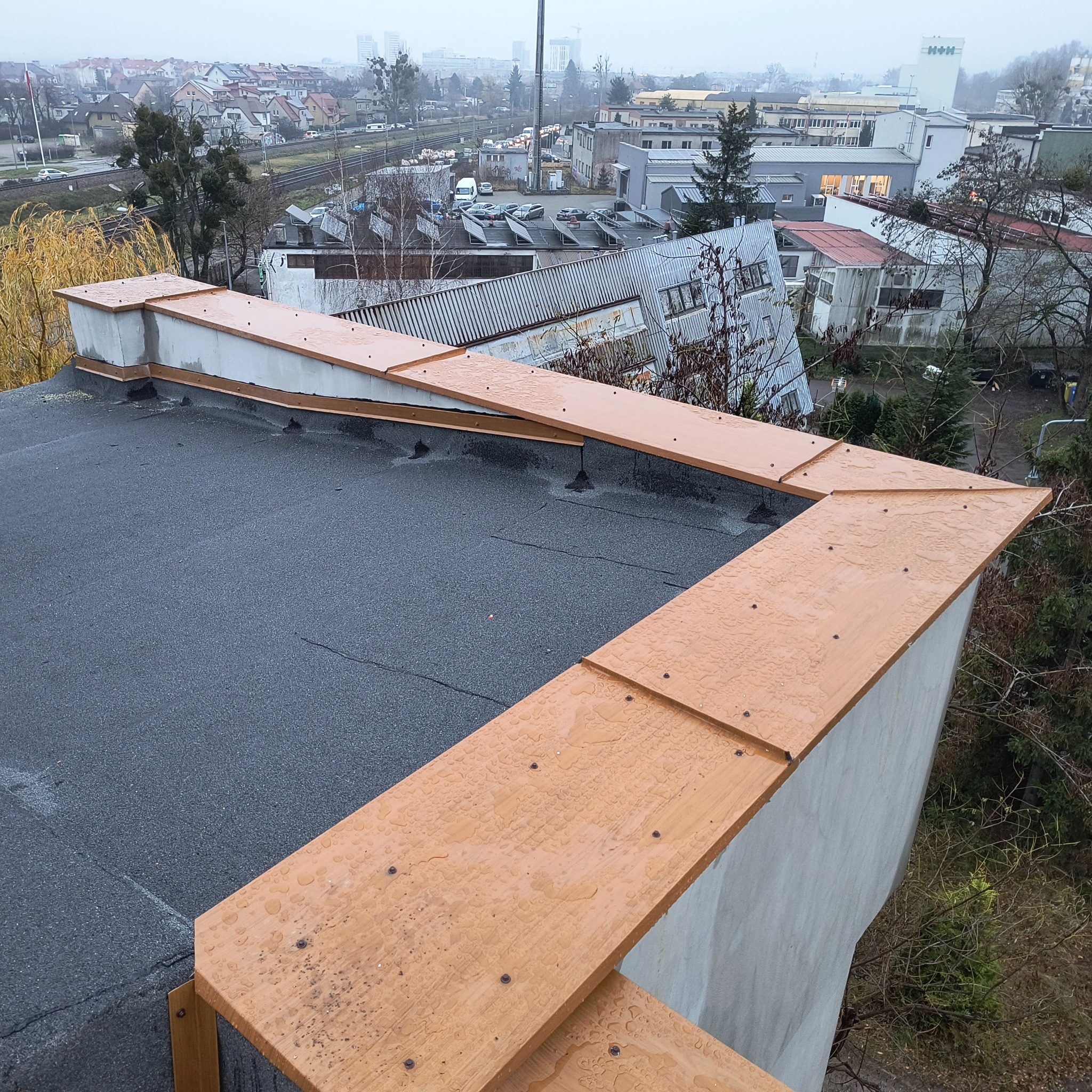 Widok z góry na fragment dachu pokrytego papą z drewnianymi obróbkami blacharskimi w kolorze jasnego brązu, na których widoczne są krople deszczu; w tle zabudowa miejska w pochmurny dzień.