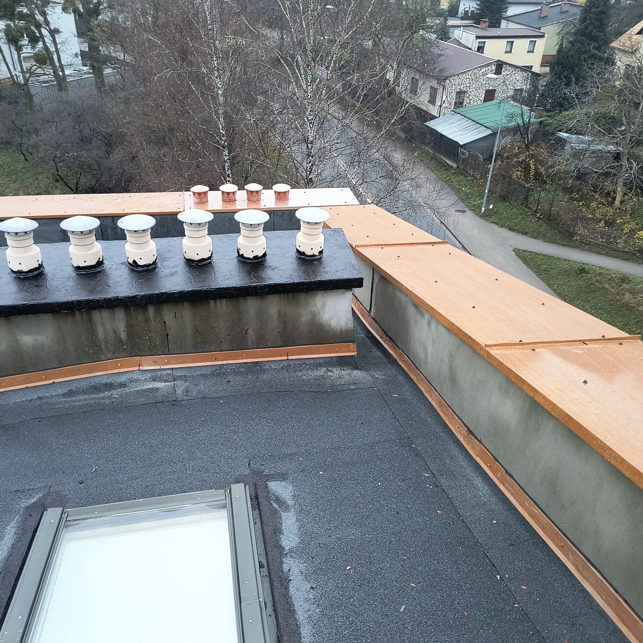 Widok z góry na płaski dach pokryty papą z lukarną i kominami wentylacyjnymi, z widocznymi elementami obróbki blacharskiej i otoczeniem miejskim w tle.