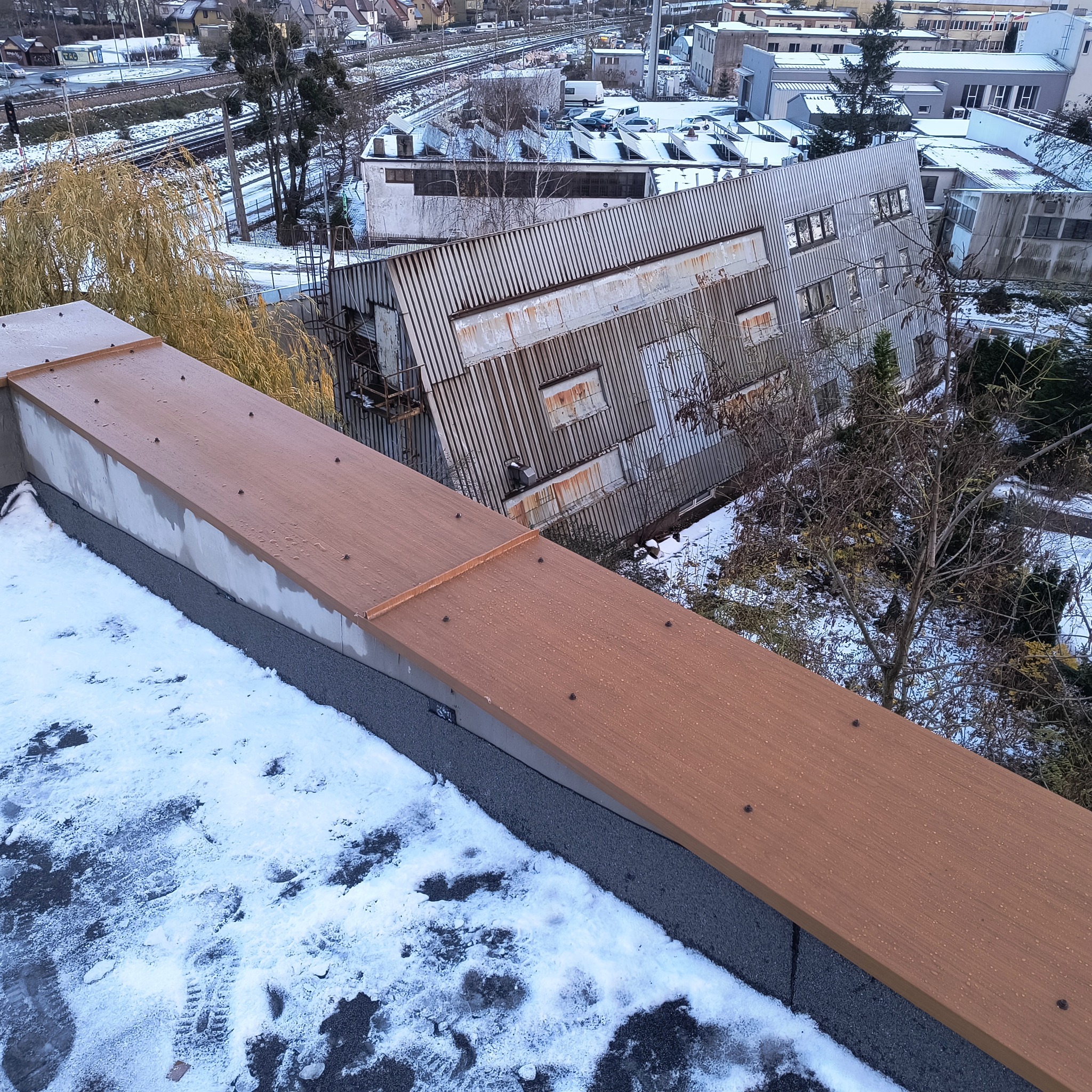 Widok z góry na nowy, brązowy dach z wykończeniem, pokryty kroplami wody po deszczu, w tle zardzewiały, blaszany dach budynku przemysłowego i śnieżny krajobraz miejski.
