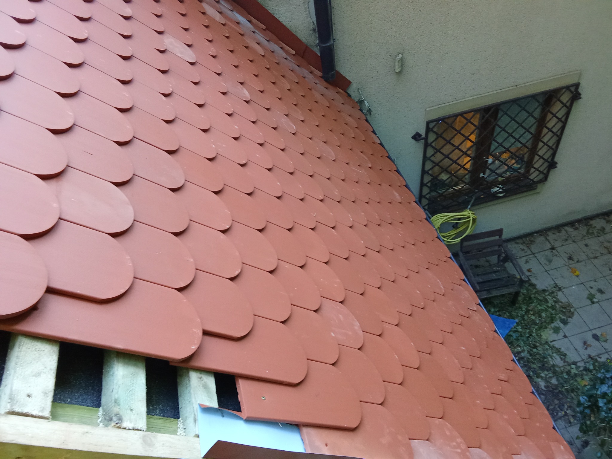 Widok z góry na fragment dachu krytego dachówką ceramiczną w kształcie łuski, z widoczną konstrukcją drewnianą i izolacją, obok ściana budynku z oknem zabezpieczonym kratą oraz zwinięty kabel...