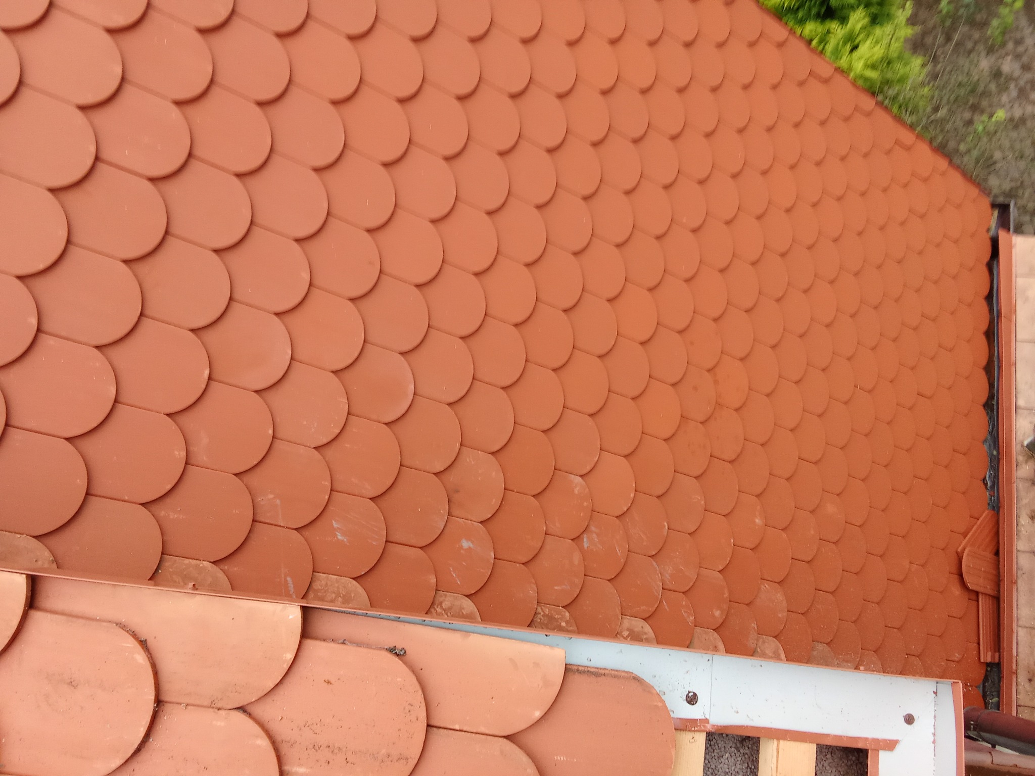Dach pokryty dachówką bitumiczną w kształcie rybiej łuski, widok z góry, z fragmentem konstrukcji dachu w dolnej części kadru i zielenią w tle.