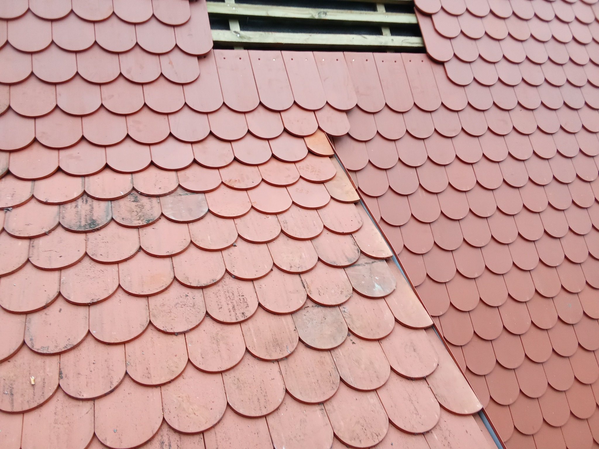 Fragment dachu z okrągłą dachówką w odcieniach czerwieni, z widocznym fragmentem konstrukcji drewnianej w miejscu ubytku.