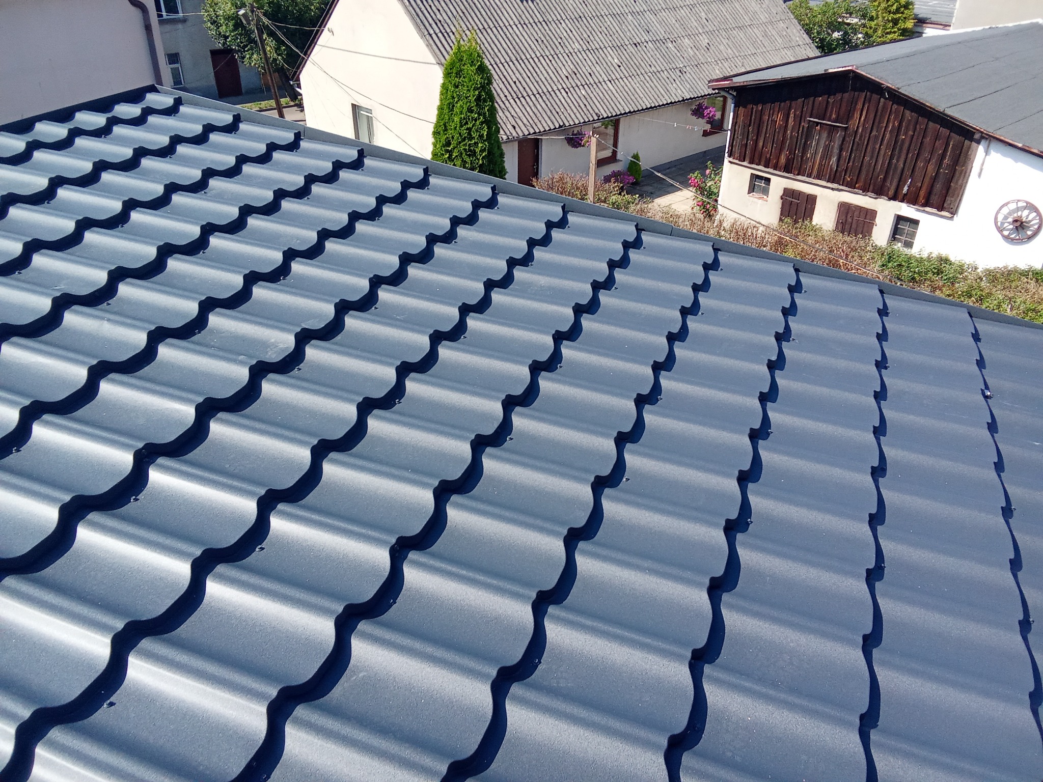 Nowy, ciemnoszary dach z blachodachówki modułowej, widok z góry na tle zabudowań mieszkalnych.