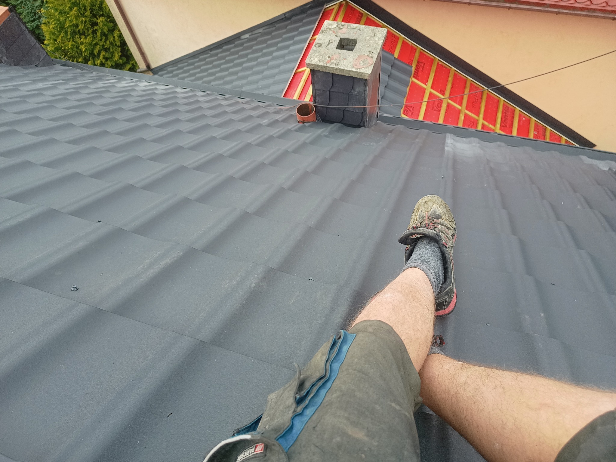 Ciemnoszary dach z blachy na etapie montażu, widoczny komin z szarych płytek, fragment czerwonej membrany dachowej oraz noga dekarza w sportowym bucie.