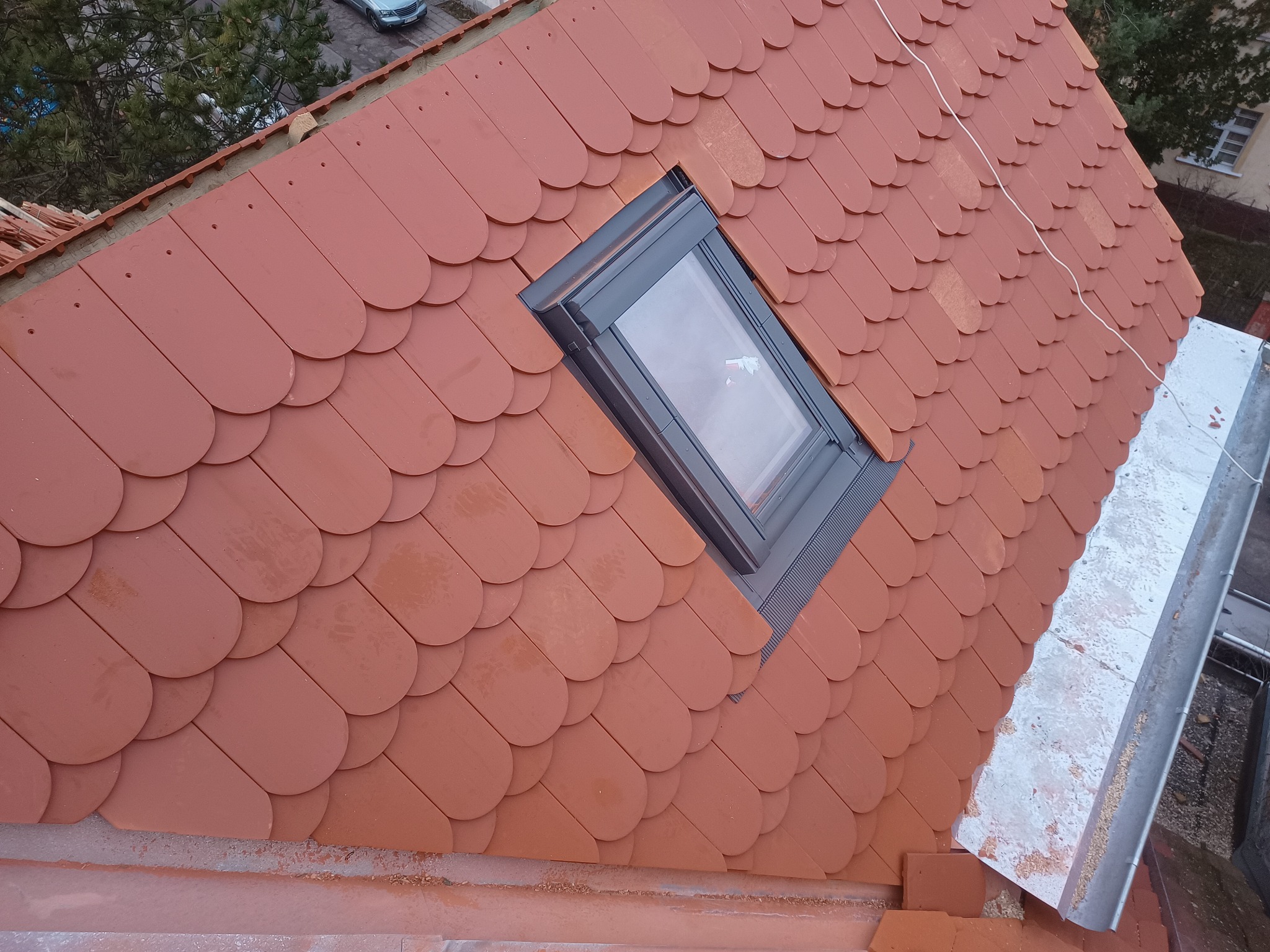 Dach pokryty czerwoną dachówką bitumiczną w kształcie łusek, z zamontowanym oknem dachowym i widoczną rynną w prawym górnym rogu.