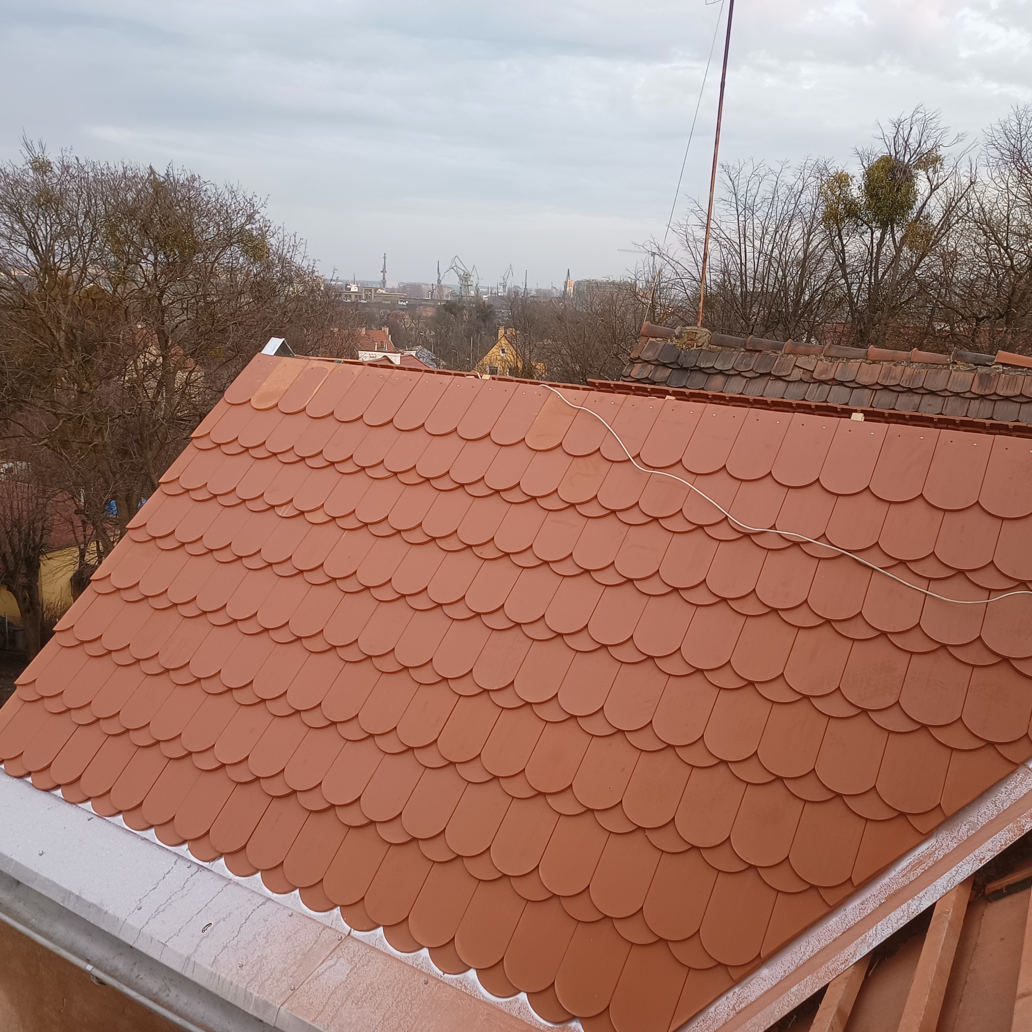 Nowy dach pokryty czerwoną dachówką karpiówką, widoczne elementy montażowe, w tle panorama miasta i drzewa bez liści.