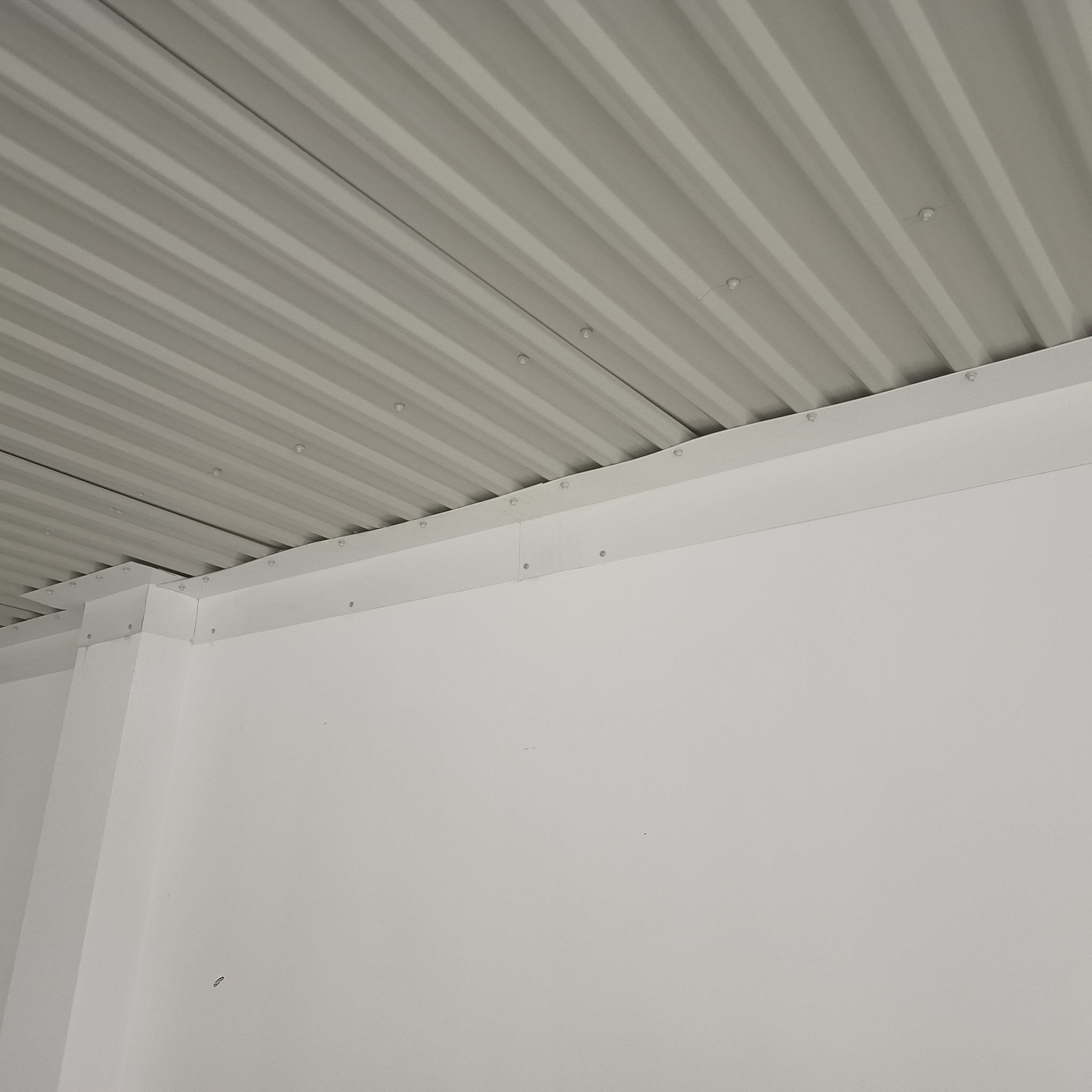 Szara blacha trapezowa tworząca zadaszenie, przymocowana do białej ściany i konstrukcji nośnej za pomocą śrub.