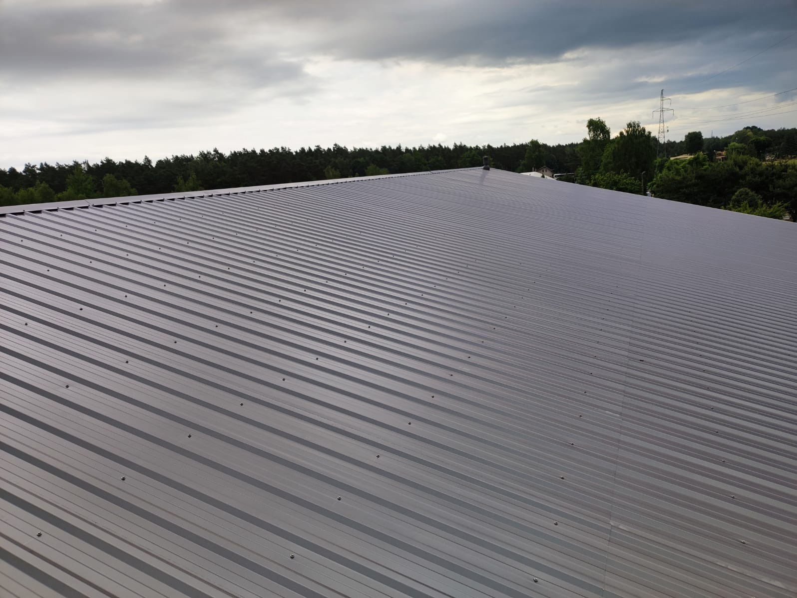 Rozległy, szary dach z blachy trapezowej, widok z góry, na tle lasu i pochmurnego nieba.