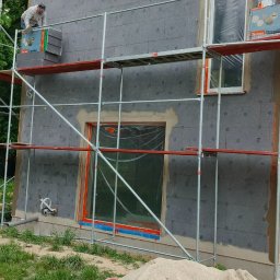 Kos-bud usługiogulno budowlane - Ocieplanie elewacji domu: pracownik na rusztowaniu montuje szare płyty termoizolacyjne. Okna zabezpieczone folią, widoczny stos materiałów budowlanych.