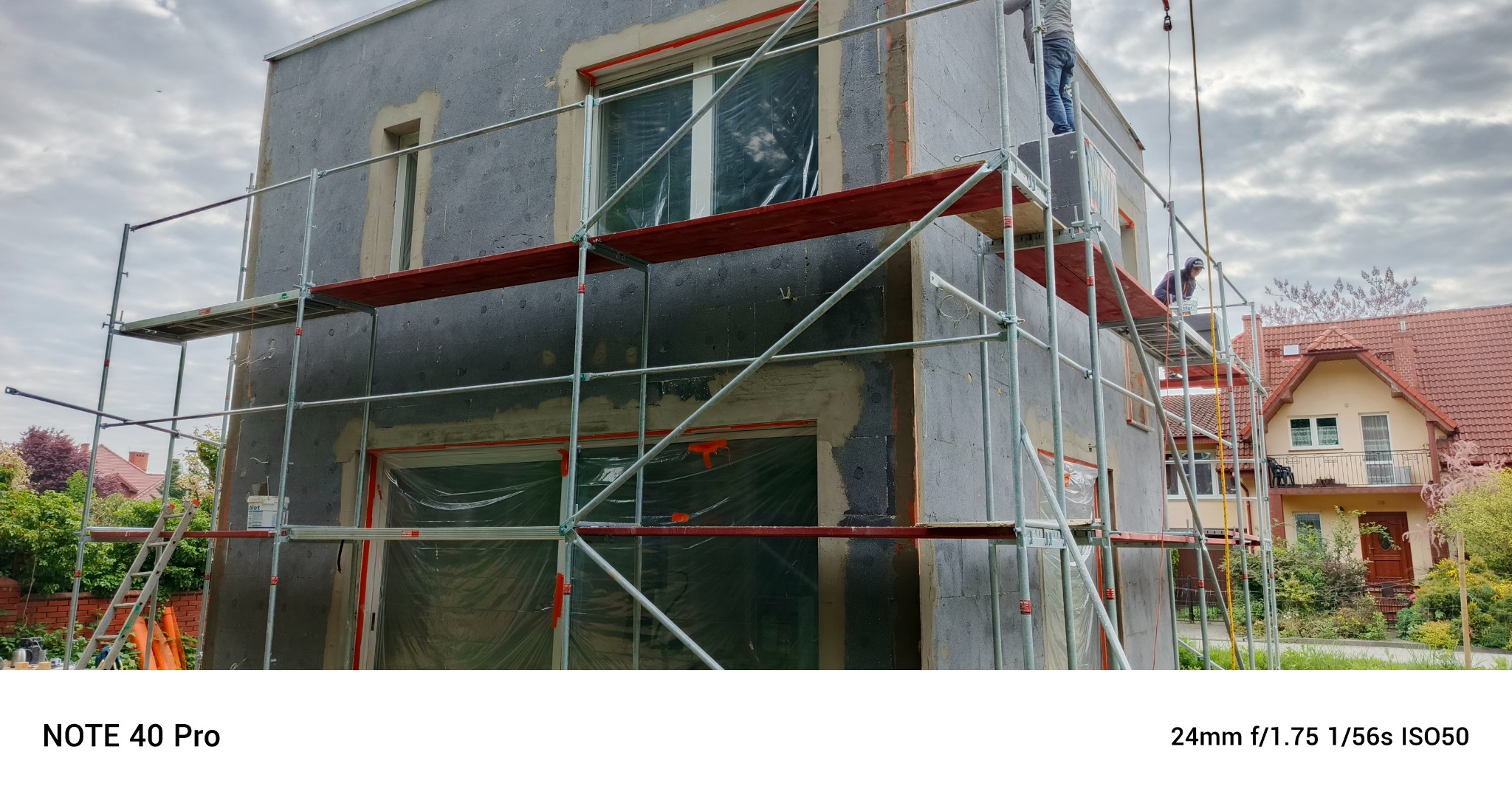 Dom w trakcie ocieplania: szare płyty izolacyjne, rusztowania, okna zabezpieczone folią. Widoczni pracownicy na rusztowaniu. Tło: dom z czerwonym dachem.