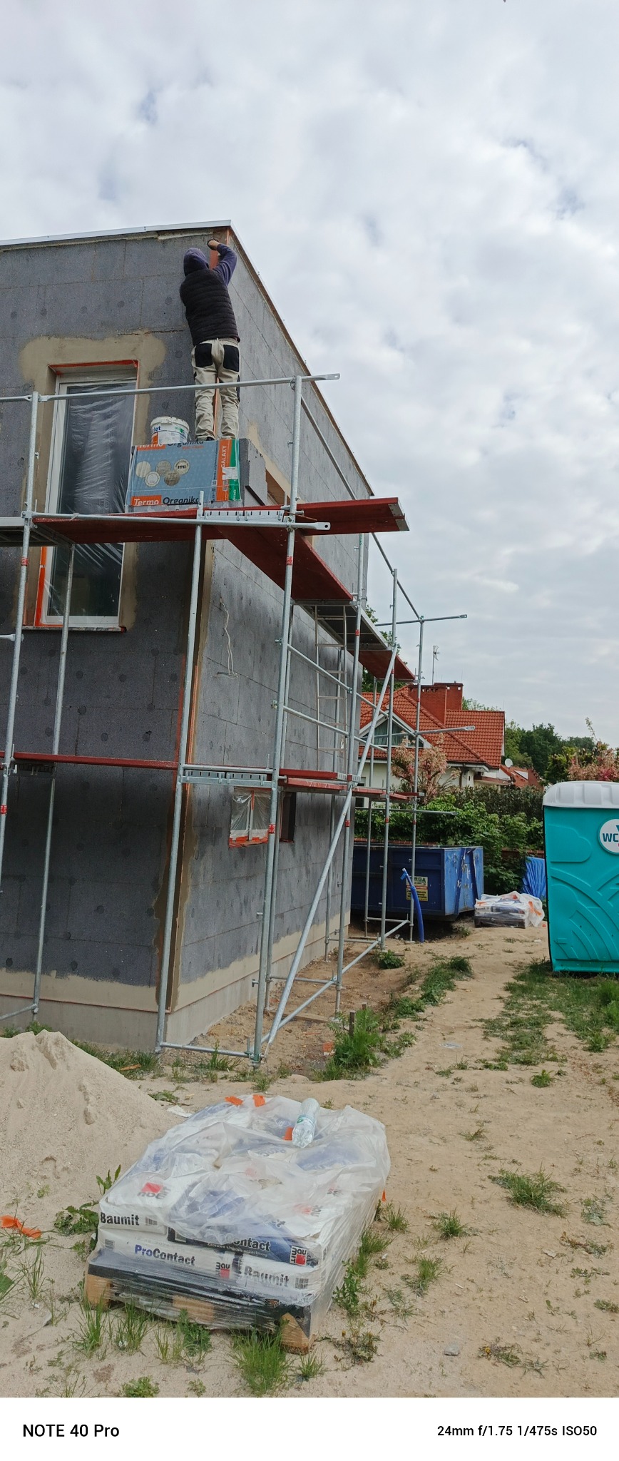 Pracownik na rusztowaniu przy ocieplonym budynku. Widoczne płyty termoizolacyjne, okno z folią, paleta z materiałami budowlanymi i przenośna toaleta w tle.