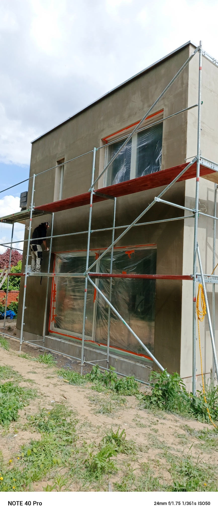 Pracownik na rusztowaniu, w trakcie nakładania jasnoszarego tynku na elewację budynku. Okna zabezpieczone folią. Widoczne narzędzia i przewód elektryczny.