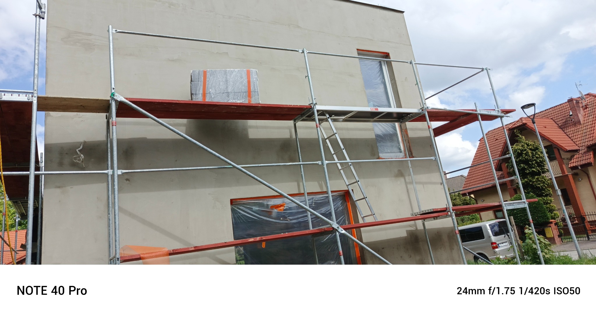 Ściana budynku w trakcie prac elewacyjnych. Widoczne rusztowania, okna zabezpieczone folią, materiał izolacyjny na podeście. Prace przygotowawcze do ocieplenia.