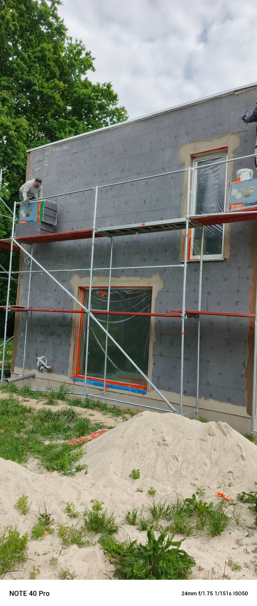 Ocieplanie elewacji domu: pracownik na rusztowaniu montuje szare płyty termoizolacyjne. Okna zabezpieczone folią, widoczny stos materiałów budowlanych.