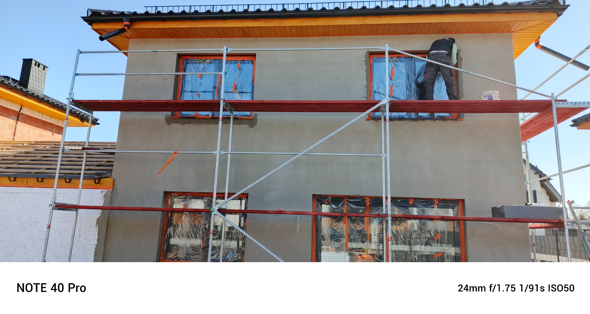 Dom w trakcie prac elewacyjnych: rusztowania, okna zabezpieczone folią, pracownik przy ścianie. Widoczny fragment ocieplonej ściany sąsiedniego budynku.