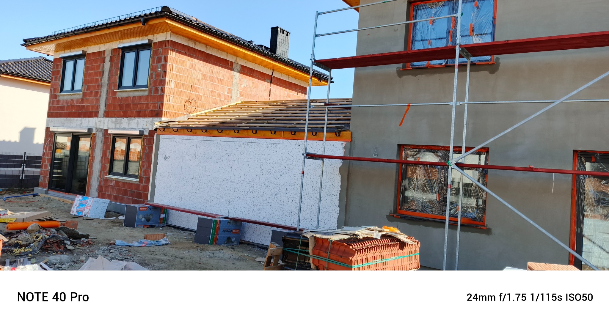 Budowa domu jednorodzinnego: ocieplona ściana z białym styropianem, surowy mur z czerwonej cegły i rusztowanie przy szarej elewacji. Widoczne materiały budowlane.