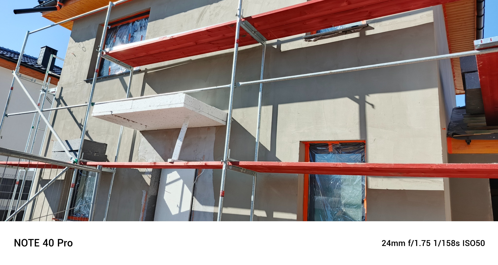 Dom w trakcie ocieplania: elewacja pokryta szarym tynkiem, rusztowania z czerwonymi deskami, okna zabezpieczone folią. Widoczny fragment ocieplenia styropianem.