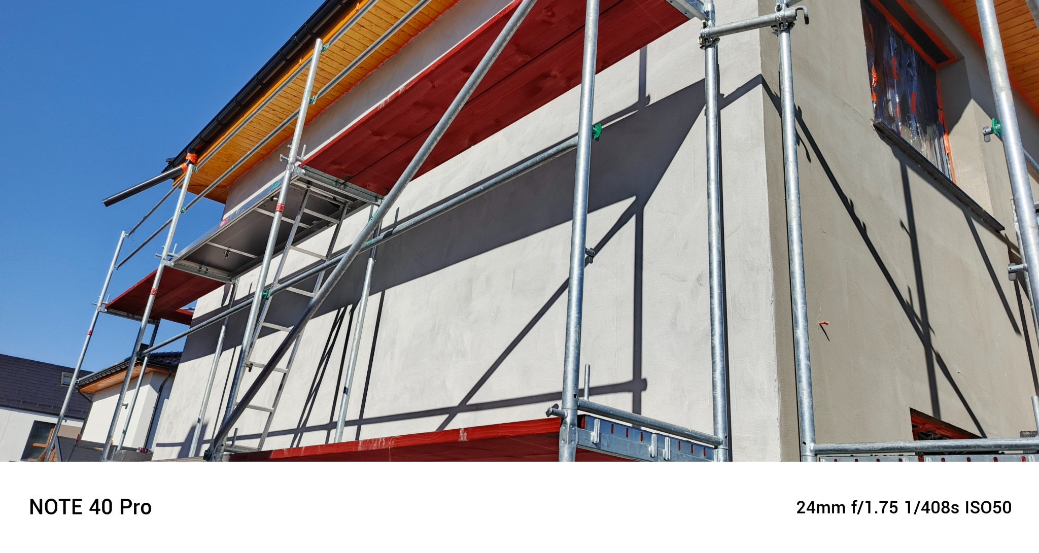 Ściana budynku w trakcie prac elewacyjnych z użyciem rusztowania. Okna zabezpieczone folią. Widoczne drewniane elementy dachu. Słoneczny dzień.