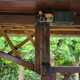 Czarna puszka elektryczna z przewodami i złączkami WAGO zamontowana na drewnianej konstrukcji altany, z widocznym włącznikiem światła poniżej.
