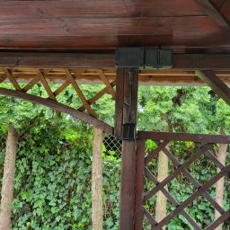 Elektryk Robert Danielak MegaWat - Zewnętrzna instalacja elektryczna w drewnianej altanie z zielonym tłem. Widoczne puszki i przewody elektryczne zamontowane na drewnianej konstrukcji.