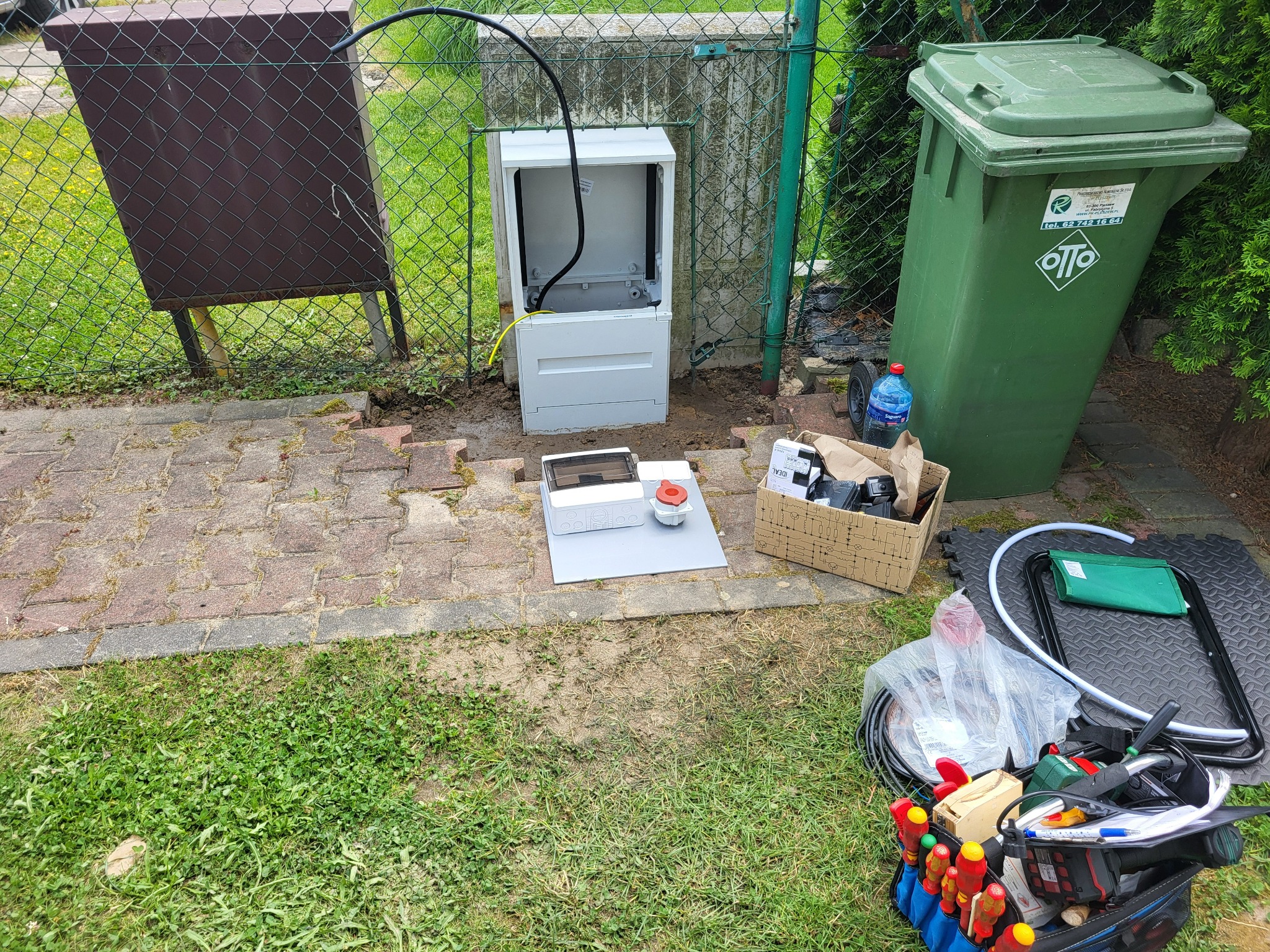 Rozdzielnica ogrodowa w trakcie montażu: gniazda 230V/400V, miejsce na sterownik podlewania. Widoczne narzędzia, kable i elementy instalacji na trawniku.