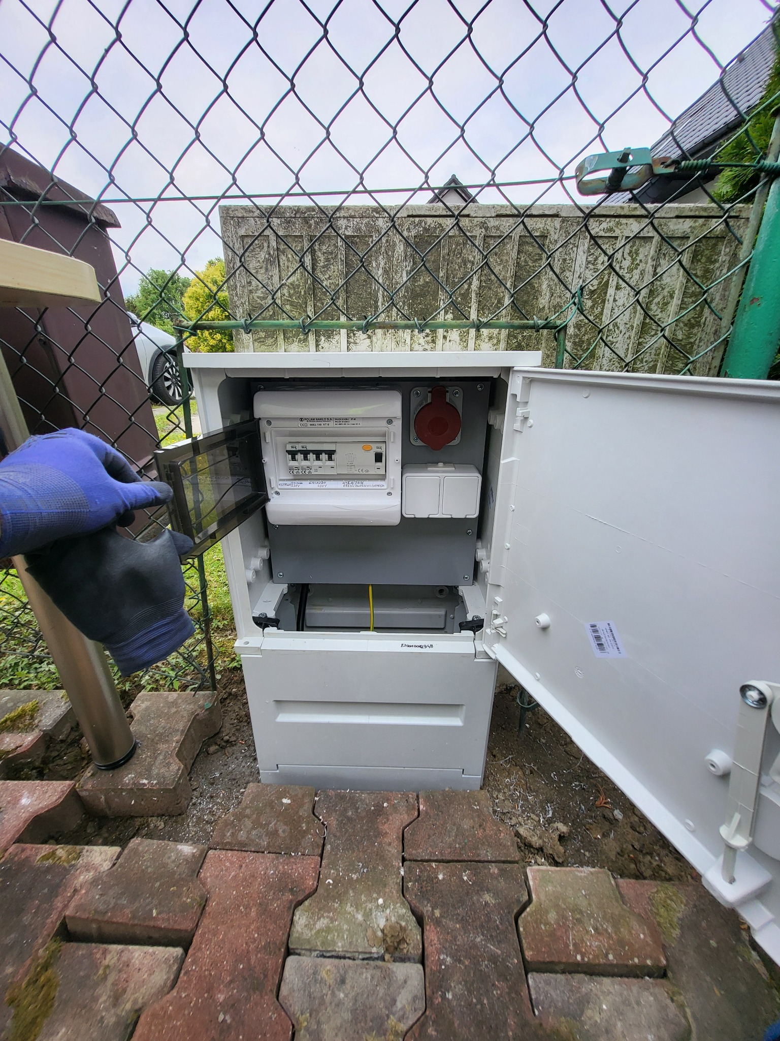 Otwarta skrzynka elektryczna z licznikiem i gniazdem, instalowana na zewnątrz, widoczna dłoń w rękawicy. Elementy instalacji w szarej obudowie na ceglanym podłożu.