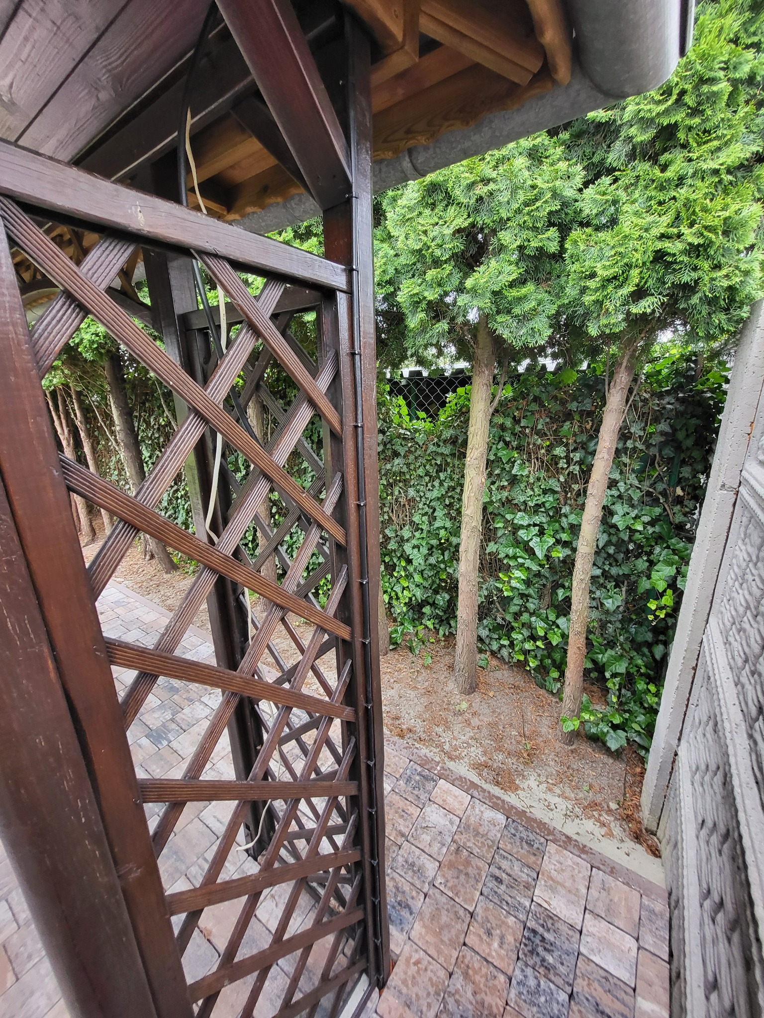 Drewniana pergola z kratką w ogrodzie, widok z dołu. Na kratce widoczne kable. Tło stanowią drzewa i kostka brukowa.
