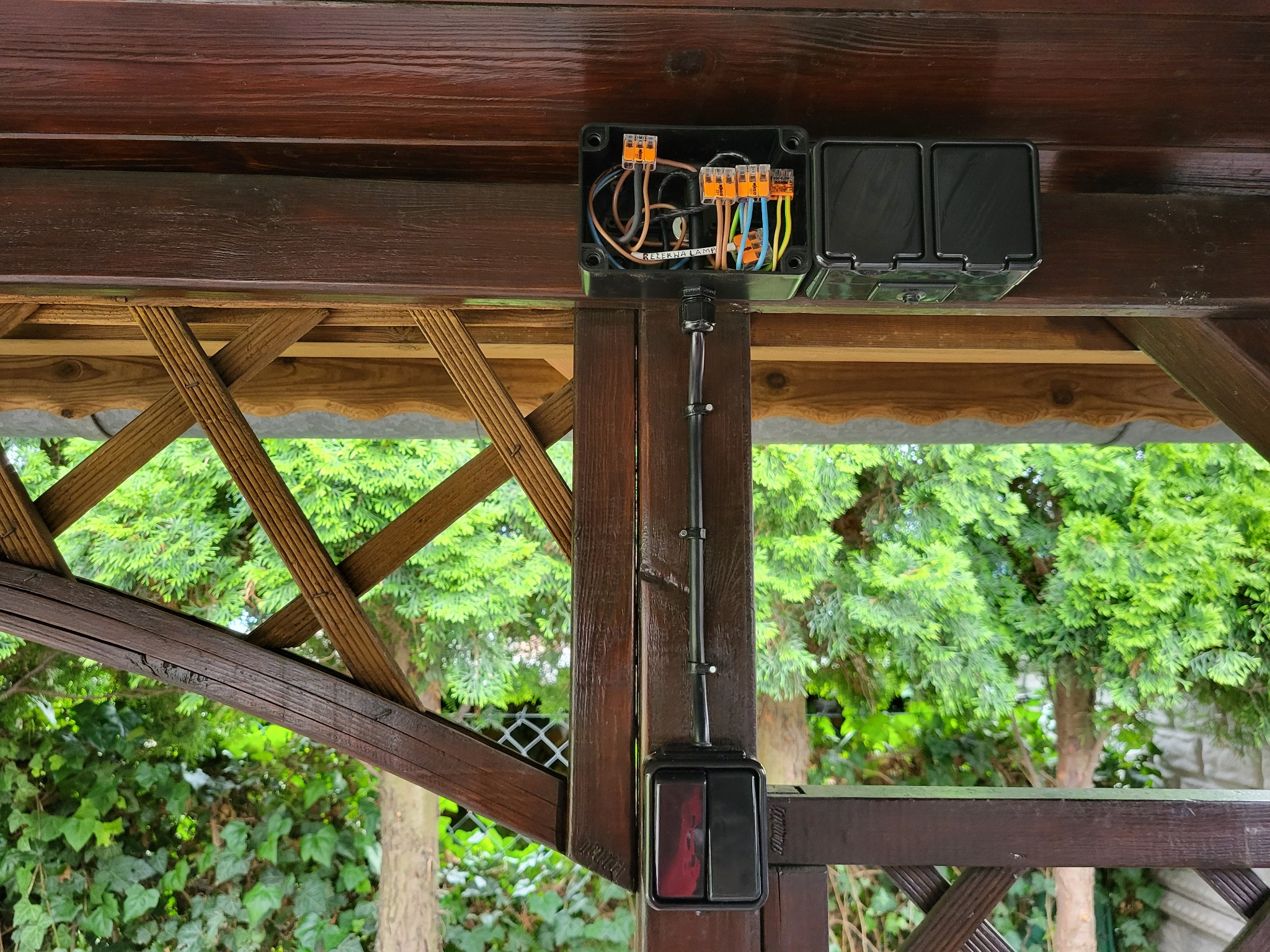 Czarna puszka elektryczna z przewodami i złączkami WAGO zamontowana na drewnianej konstrukcji altany, z widocznym włącznikiem światła poniżej.