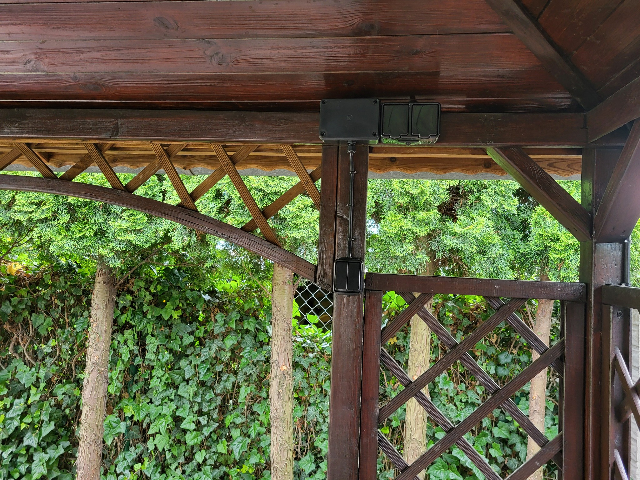Zewnętrzna instalacja elektryczna w drewnianej altanie z zielonym tłem. Widoczne puszki i przewody elektryczne zamontowane na drewnianej konstrukcji.