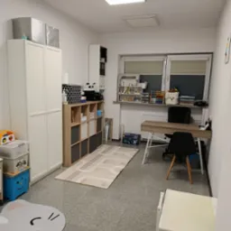 Pokój terapeutyczny z biurkiem, regałami i dywanem z motywem ulicy. Stonowana kolorystyka, jasne oświetlenie, przytulna aranżacja przestrzeni dla dzieci.