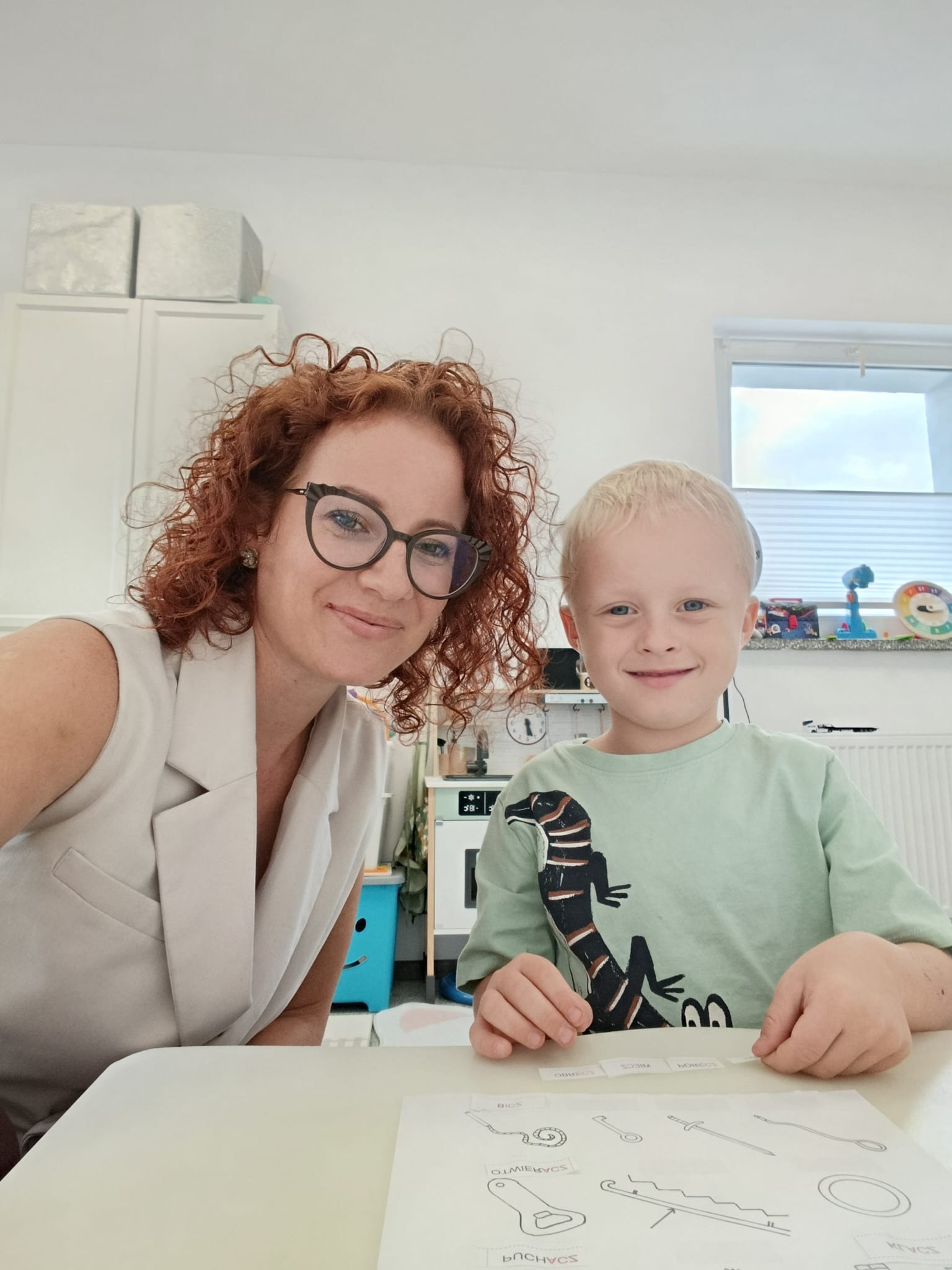 Uśmiechnięta logopedka z rudymi włosami i chłopiec z blond włosami podczas ćwiczeń logopedycznych przy stole z kartą pracy i podpisanymi obrazkami.