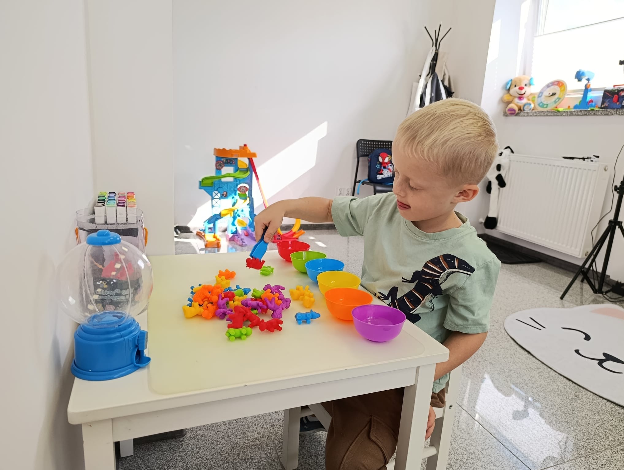 Chłopiec ćwiczy motorykę małą, używając szczypiec do przenoszenia kolorowych figurek do misek. Terapia logopedyczna z zabawkami edukacyjnymi na stole.