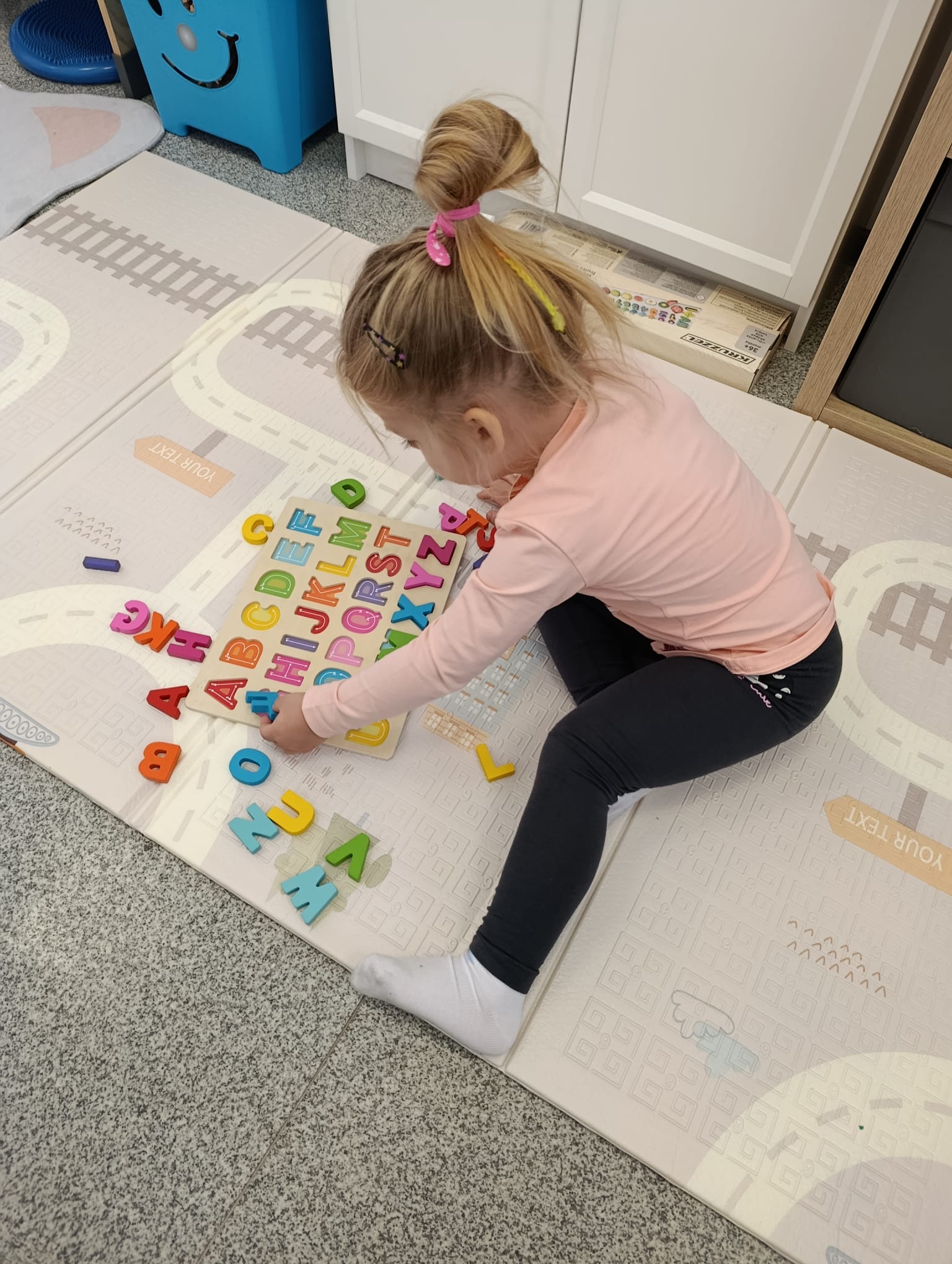 Dziewczynka układa kolorowe, drewniane literki w drewnianej ramce z alfabetem na macie edukacyjnej z motywem ulic. Zabawka logopedyczna.