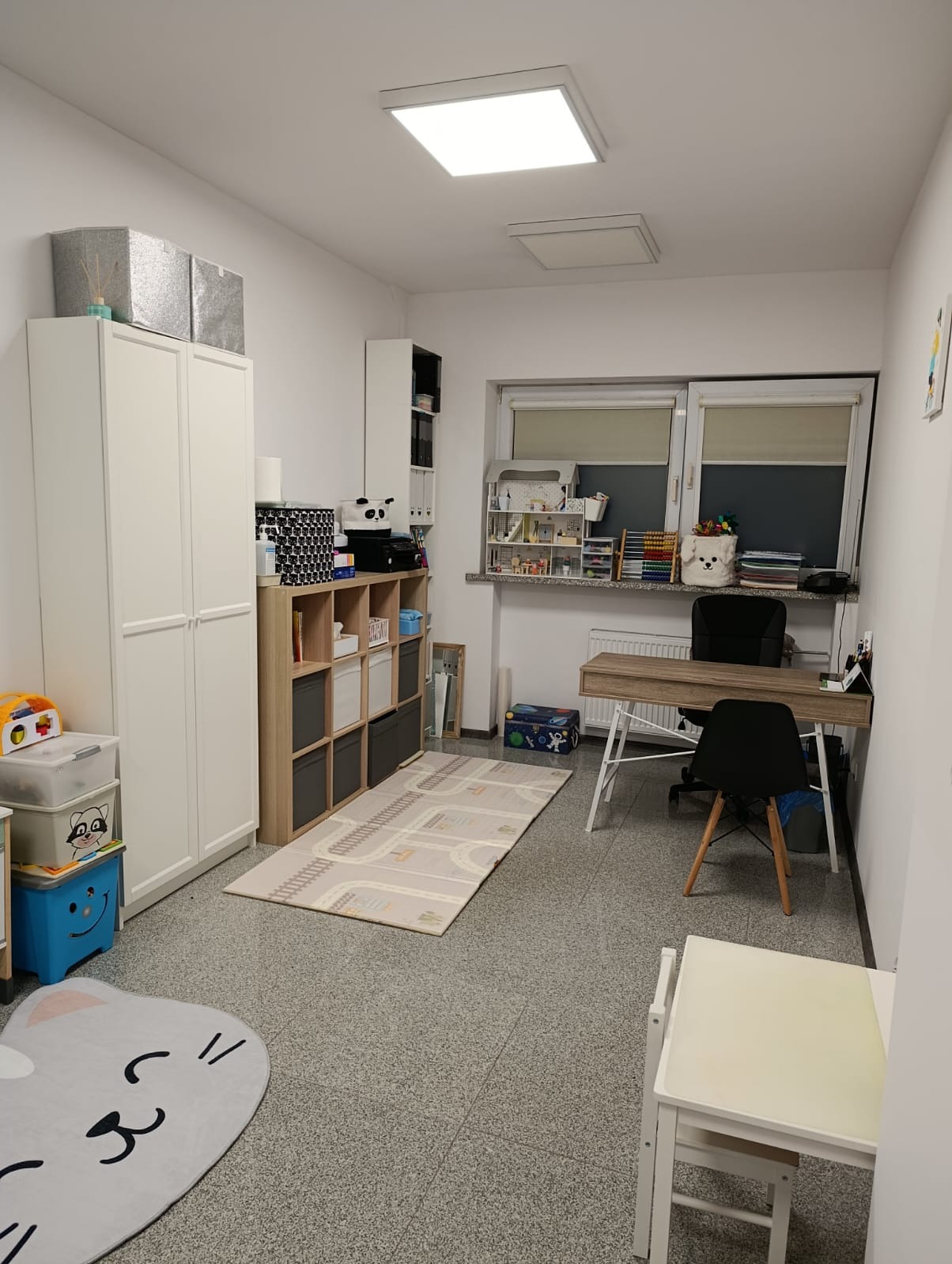 Pokój terapeutyczny z biurkiem, regałami i dywanem z motywem ulicy. Stonowana kolorystyka, jasne oświetlenie, przytulna aranżacja przestrzeni dla dzieci.