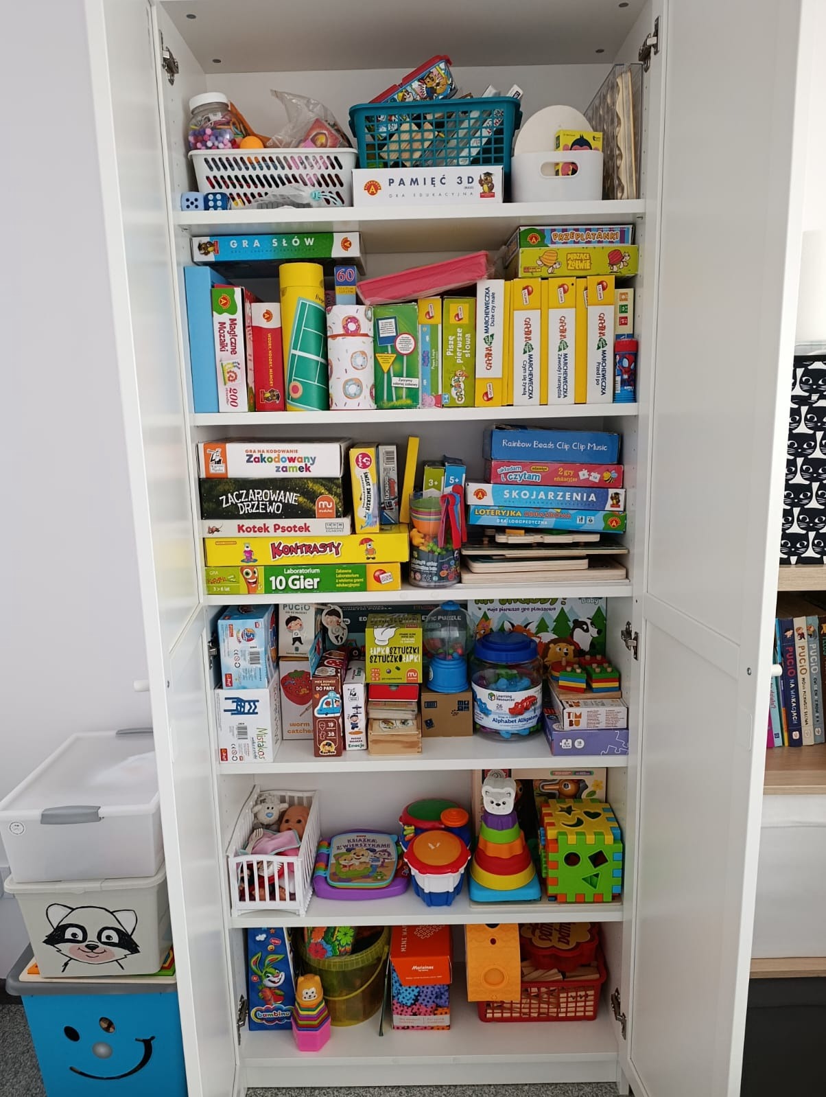 Pełna szafa z grami edukacyjnymi i zabawkami dla dzieci. Różnorodne pudełka, układanki i akcesoria logopedyczne. Białe półki, kolorowe gry.