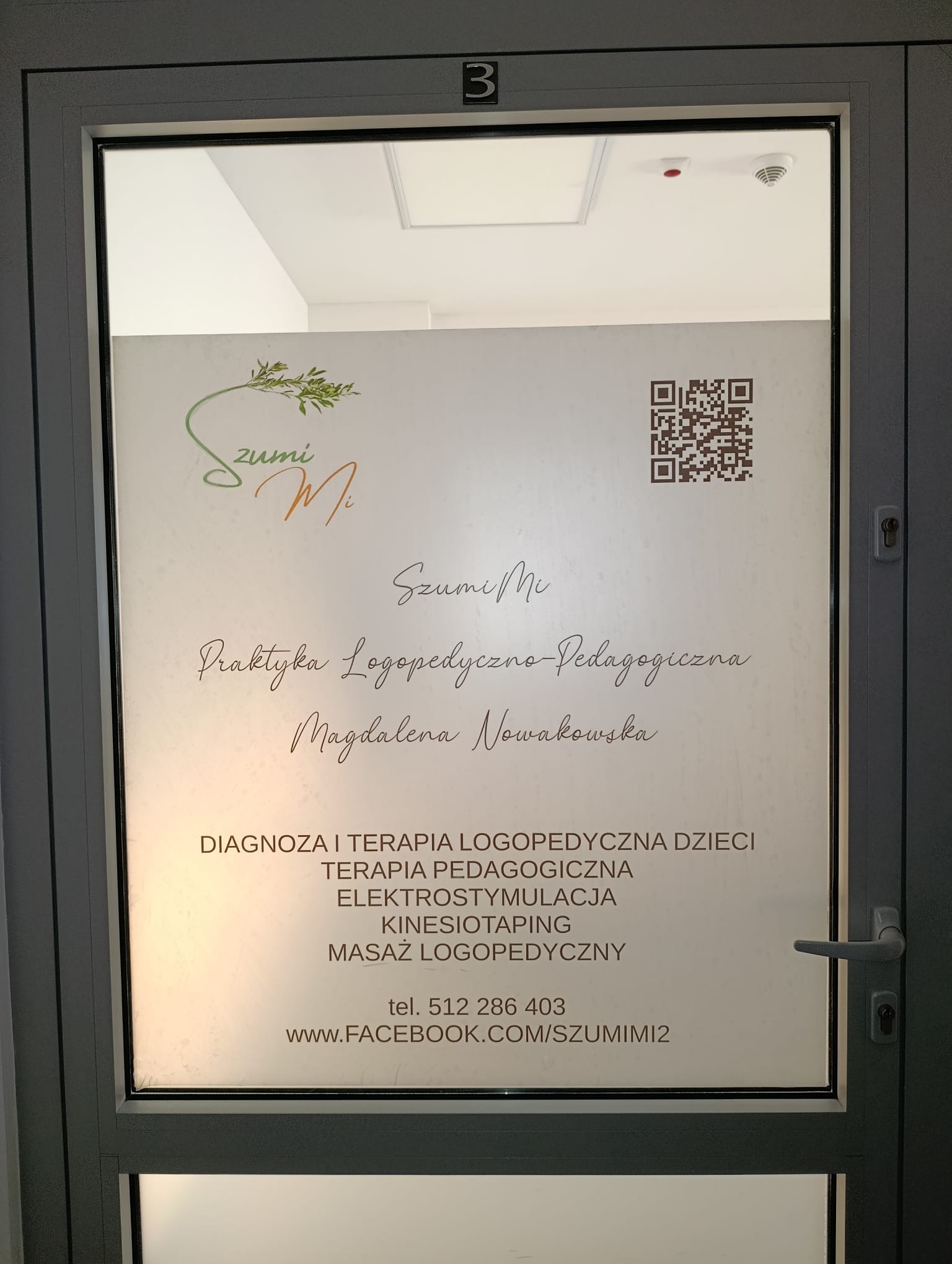 Szklane drzwi do gabinetu logopedycznego 'Szumi Mi' z informacjami o ofercie, numerem telefonu i QR kodem do strony na Facebooku. Widoczny numer '3' nad drzwiami.