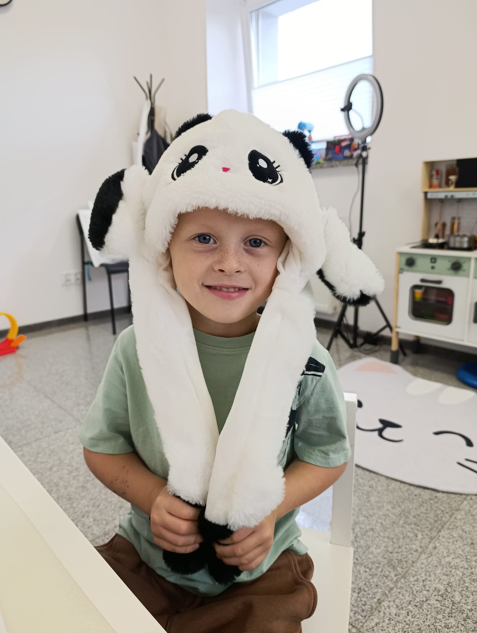 Uśmiechnięty chłopiec z piegami w pluszowej czapce pandy, w tle zabawki i lampa pierścieniowa. Sesja w gabinecie terapeutycznym.