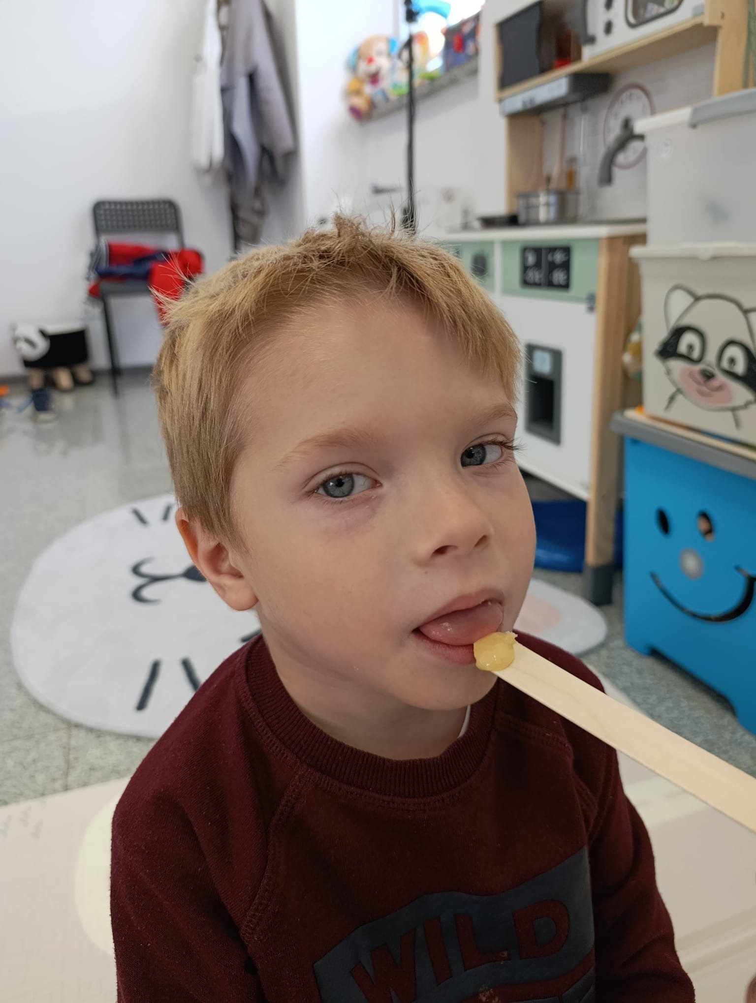 Chłopiec z blond włosami podczas badania jamy ustnej patyczkiem w gabinecie, w tle zabawki. Uwagę przykuwa skierowany do kamery wzrok dziecka.